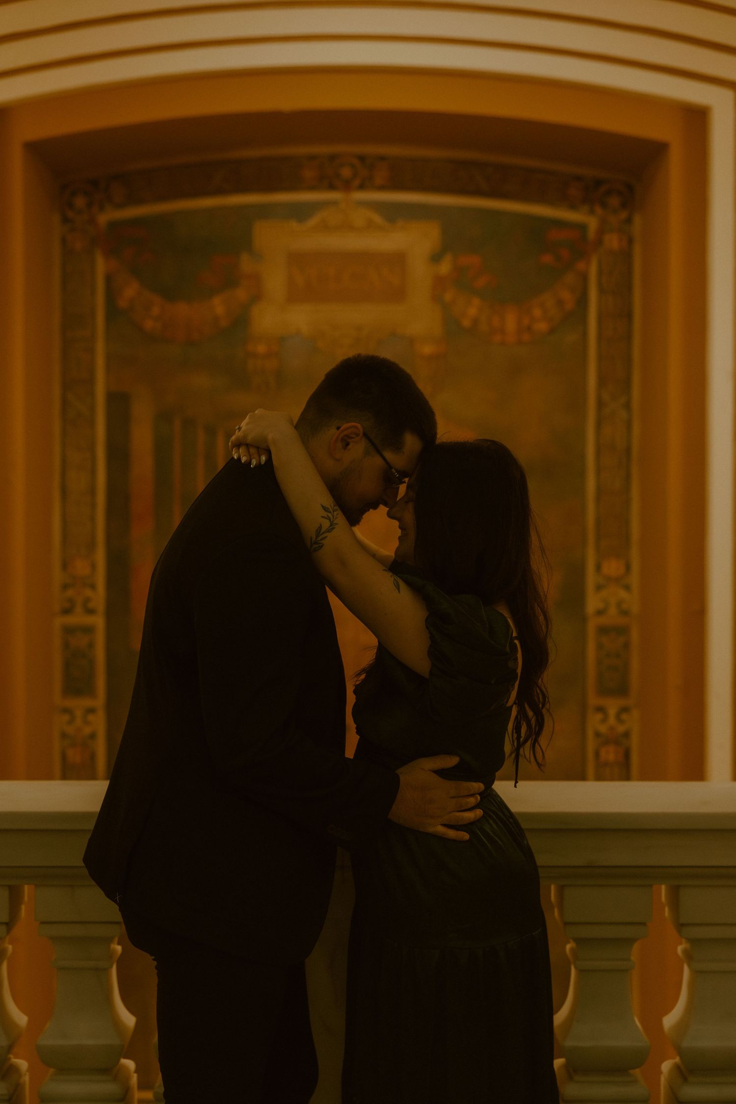 A warm-toned romantic photograph of a couple embracing in front of an ornate vintage wall with decorative details.