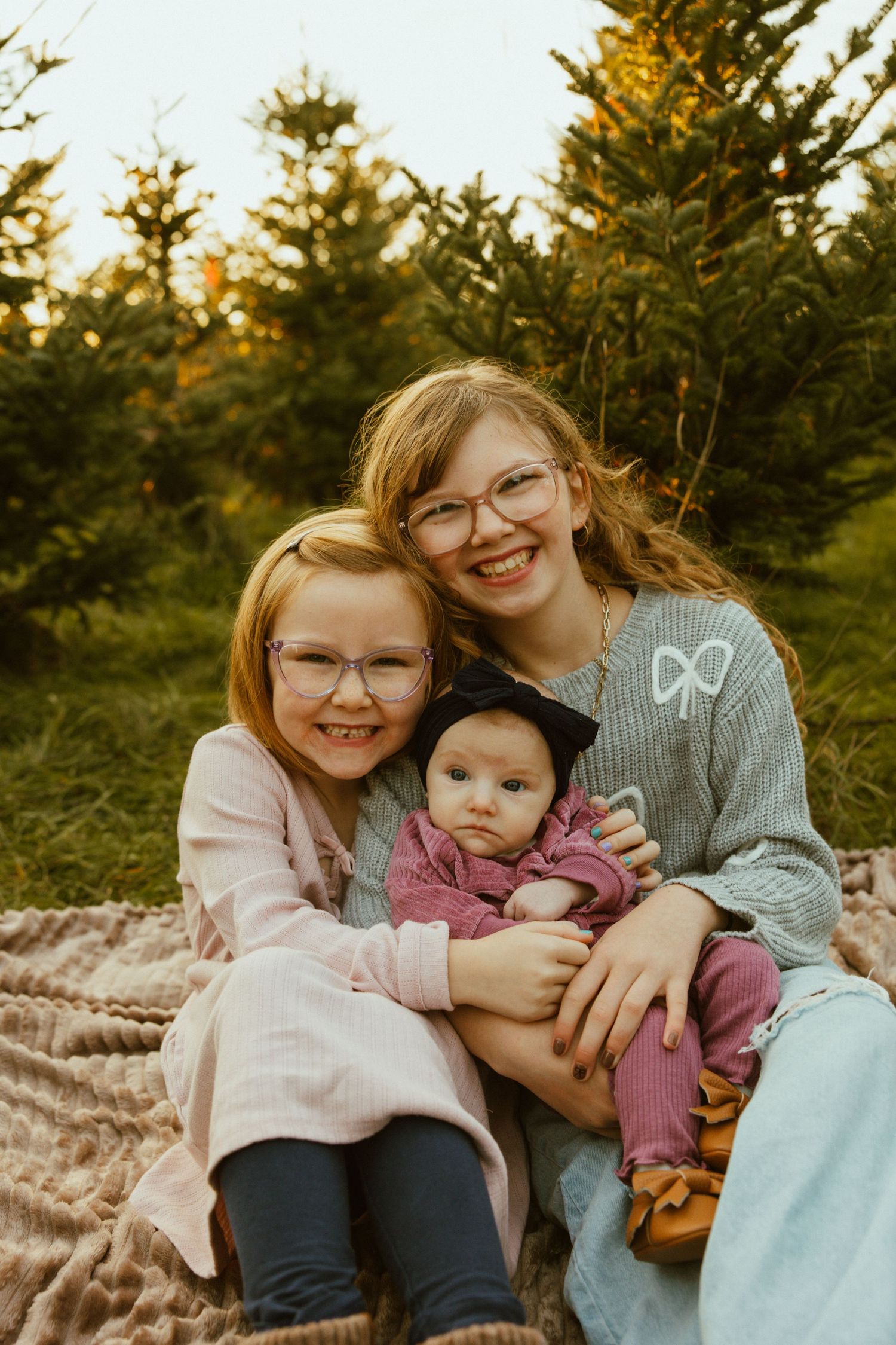 Three people sitting together on a brown blanket among evergreen trees at sunset.