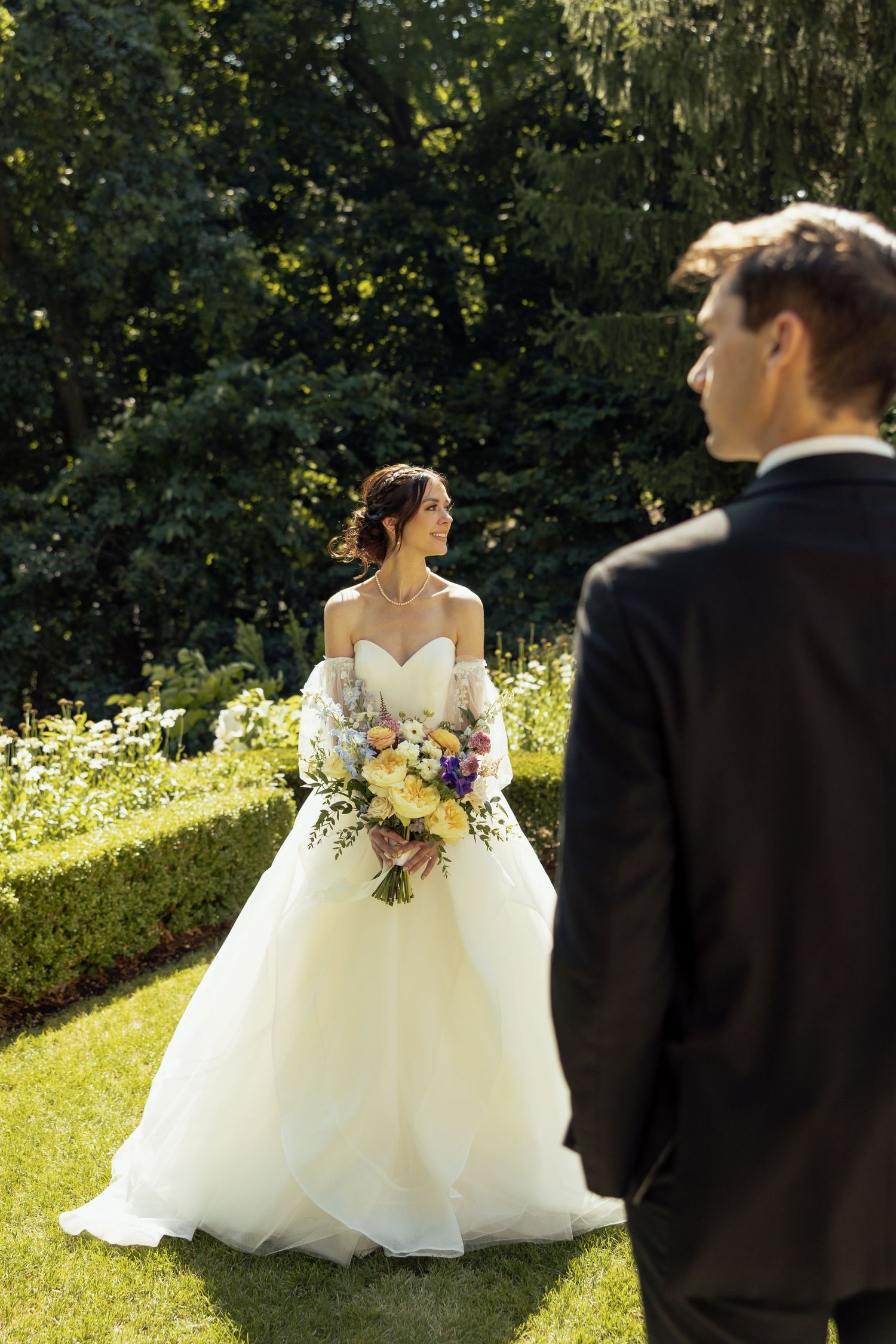 A bride in a flowing white gown poses gracefully on a sunlit garden lawn.