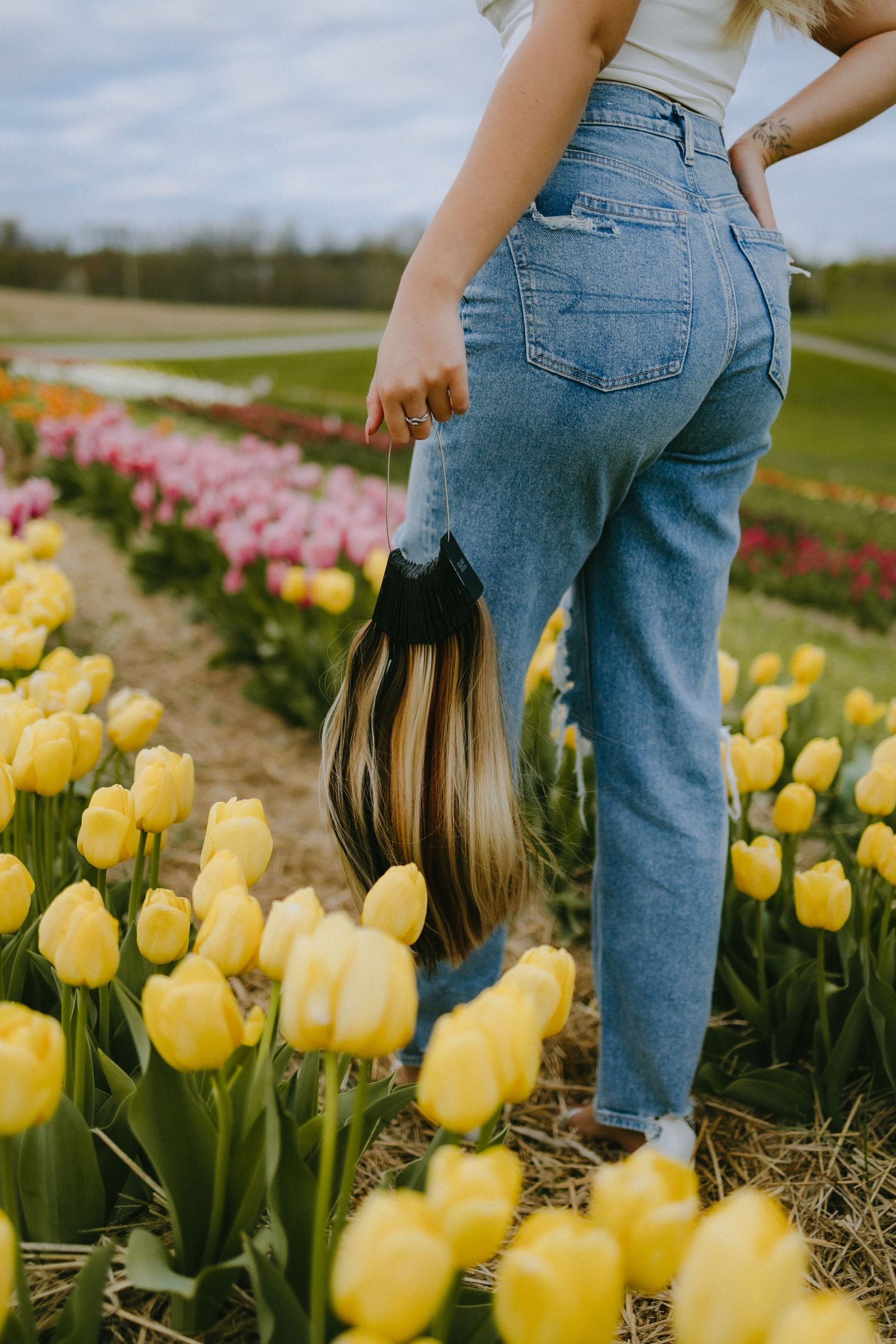 Person in blue jeans standing among yellow and pink tulip fields holding handbag.