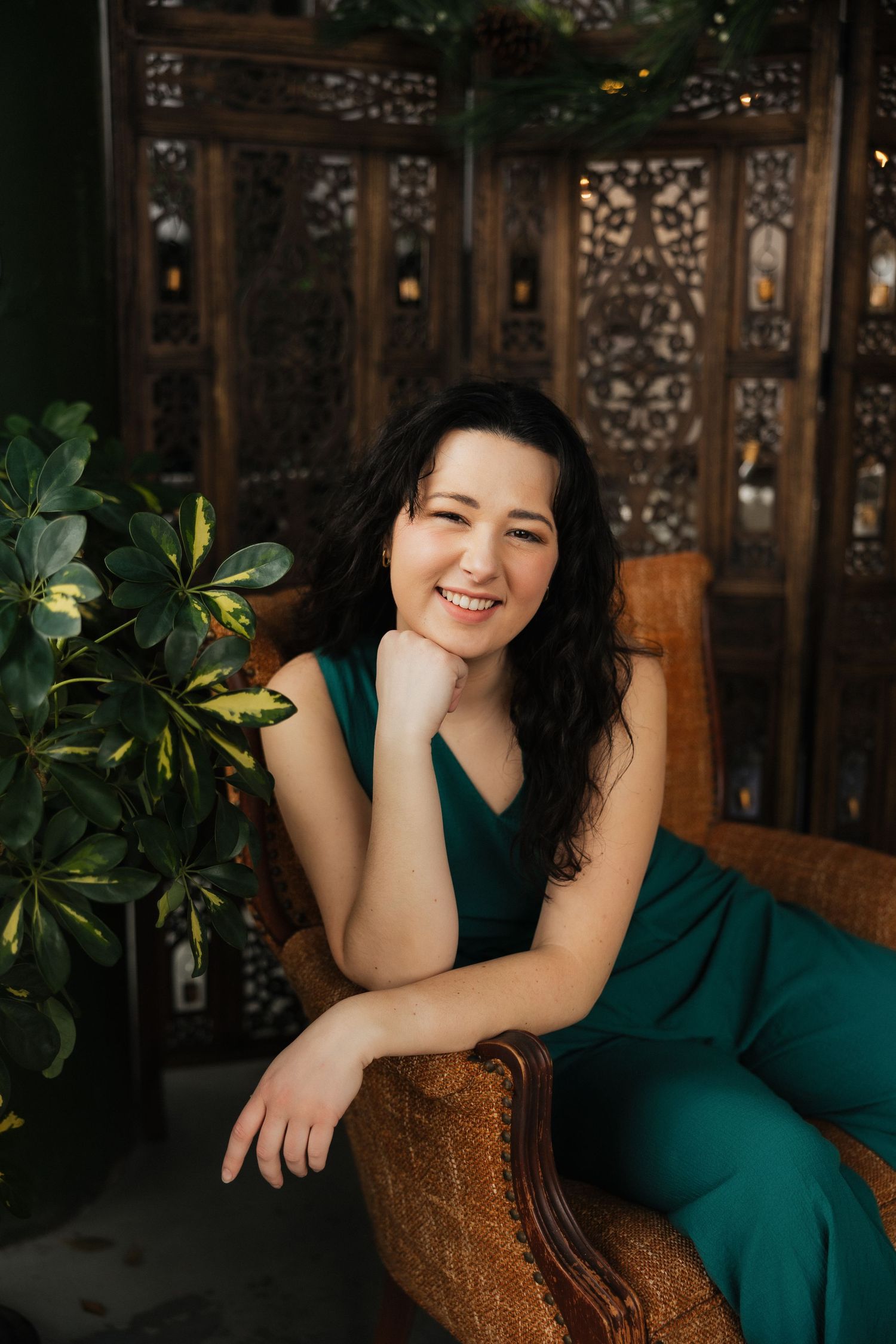 Portrait of person in green outfit seated on brown chair with decorative wall backdrop.