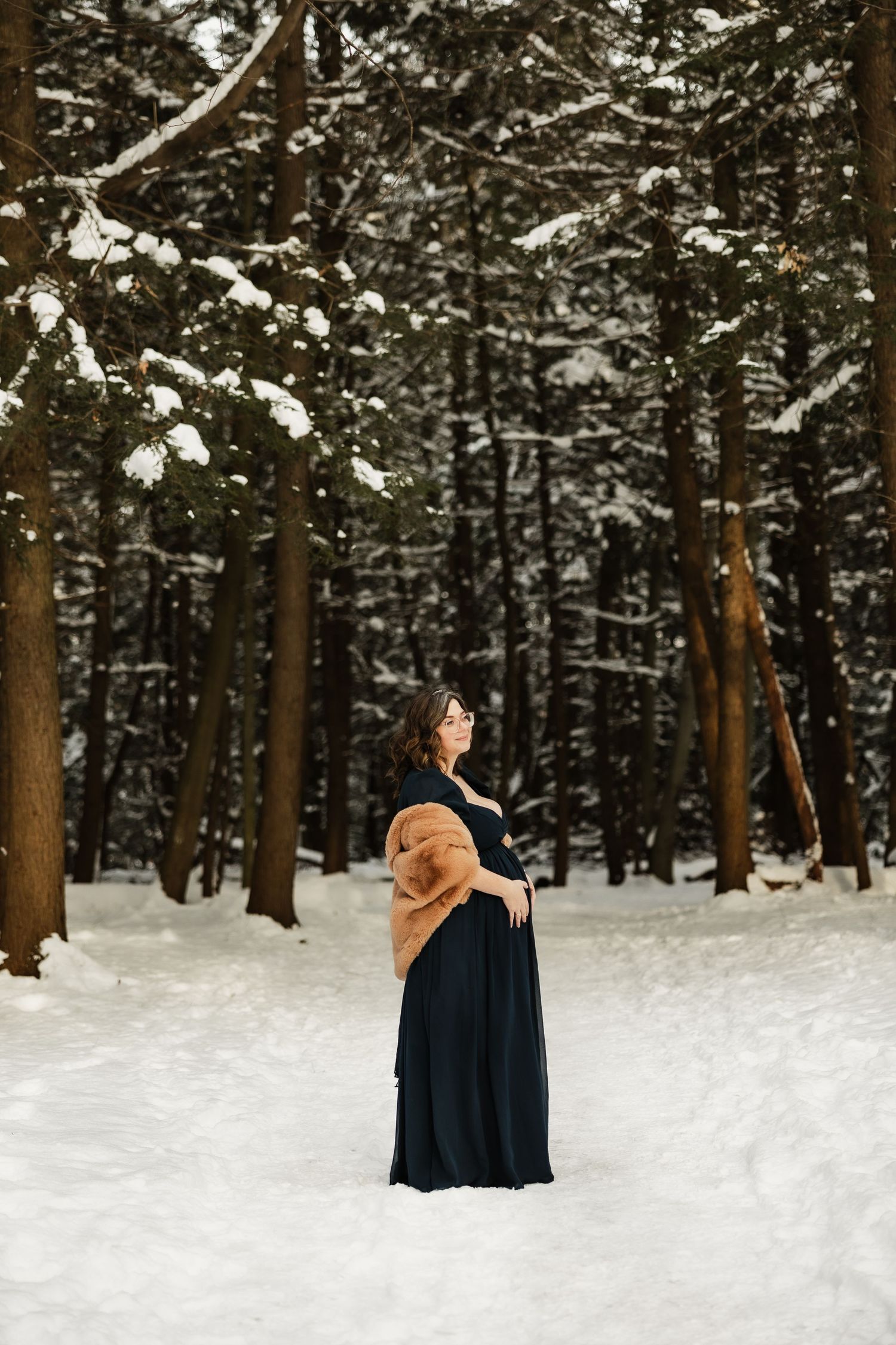 A person in a black gown and brown fur shawl stands elegantly in a snowy forest setting.