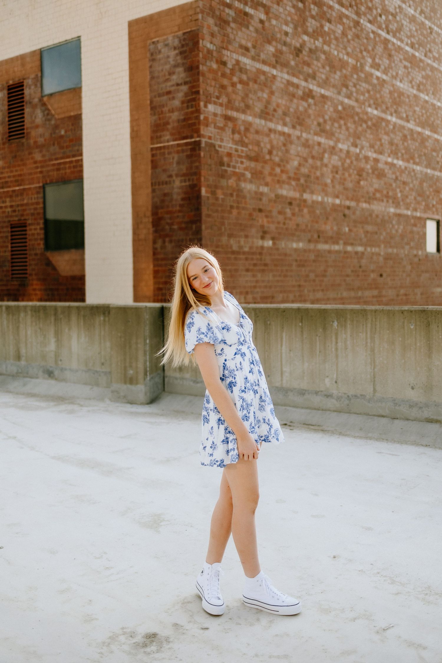 Young woman in floral blue dress and white sneakers poses against brick wall.