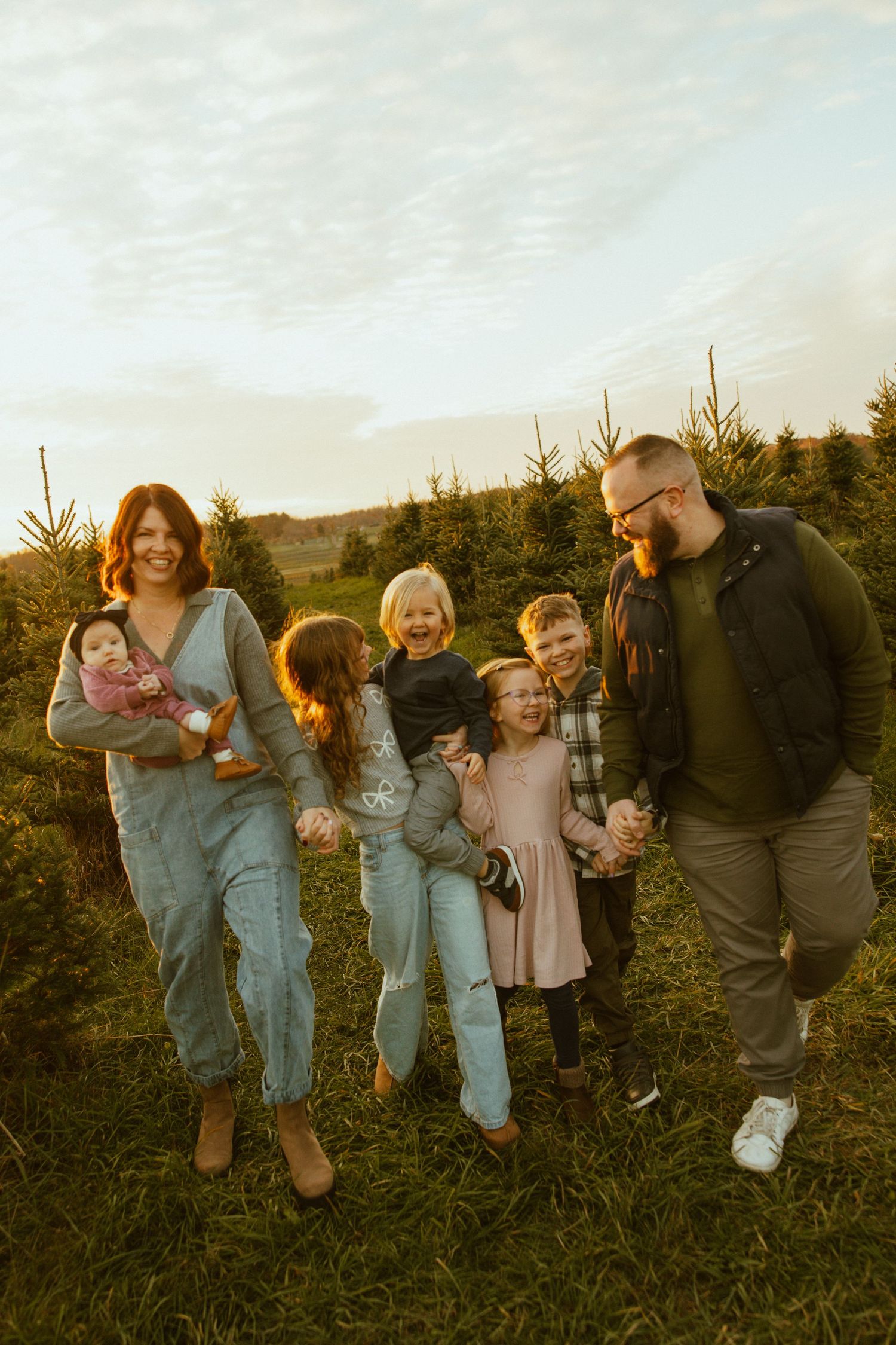 A family laughs together while standing between rows of pine trees during golden hour at a Christmas tree farm.