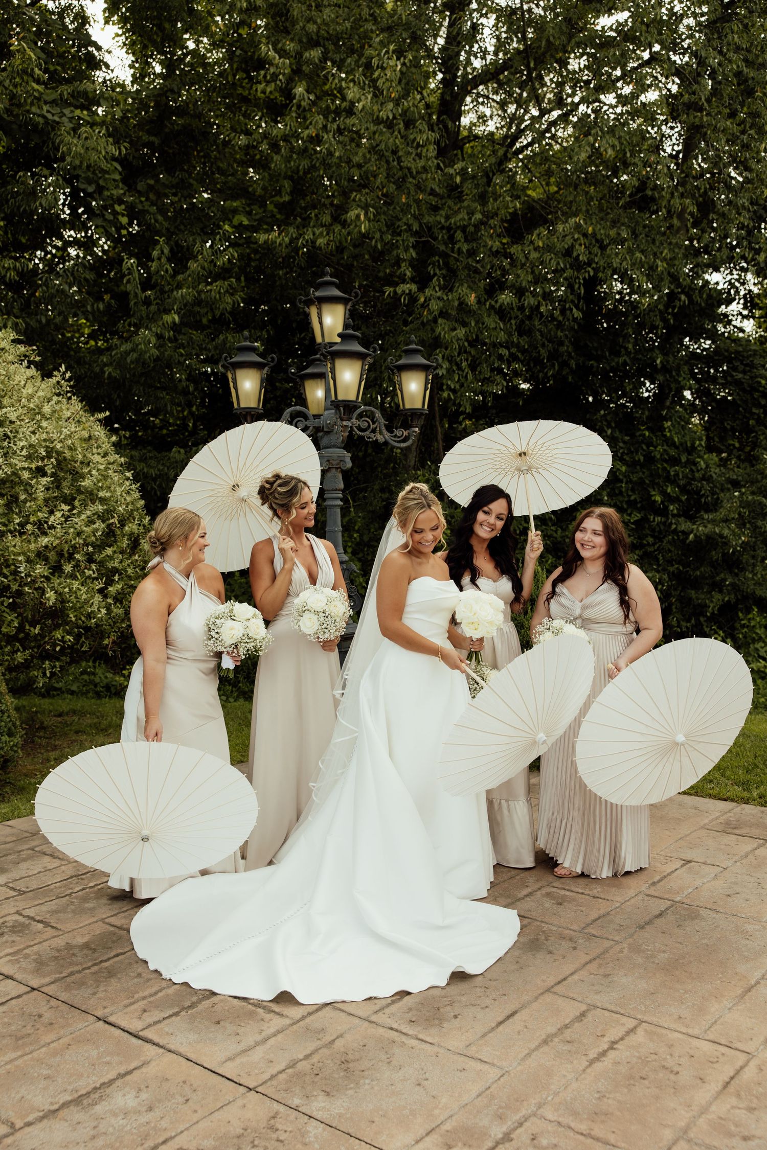 Bridesmaids in light colored dresses pose with white parasols on a stone patio during an elegant wedding.