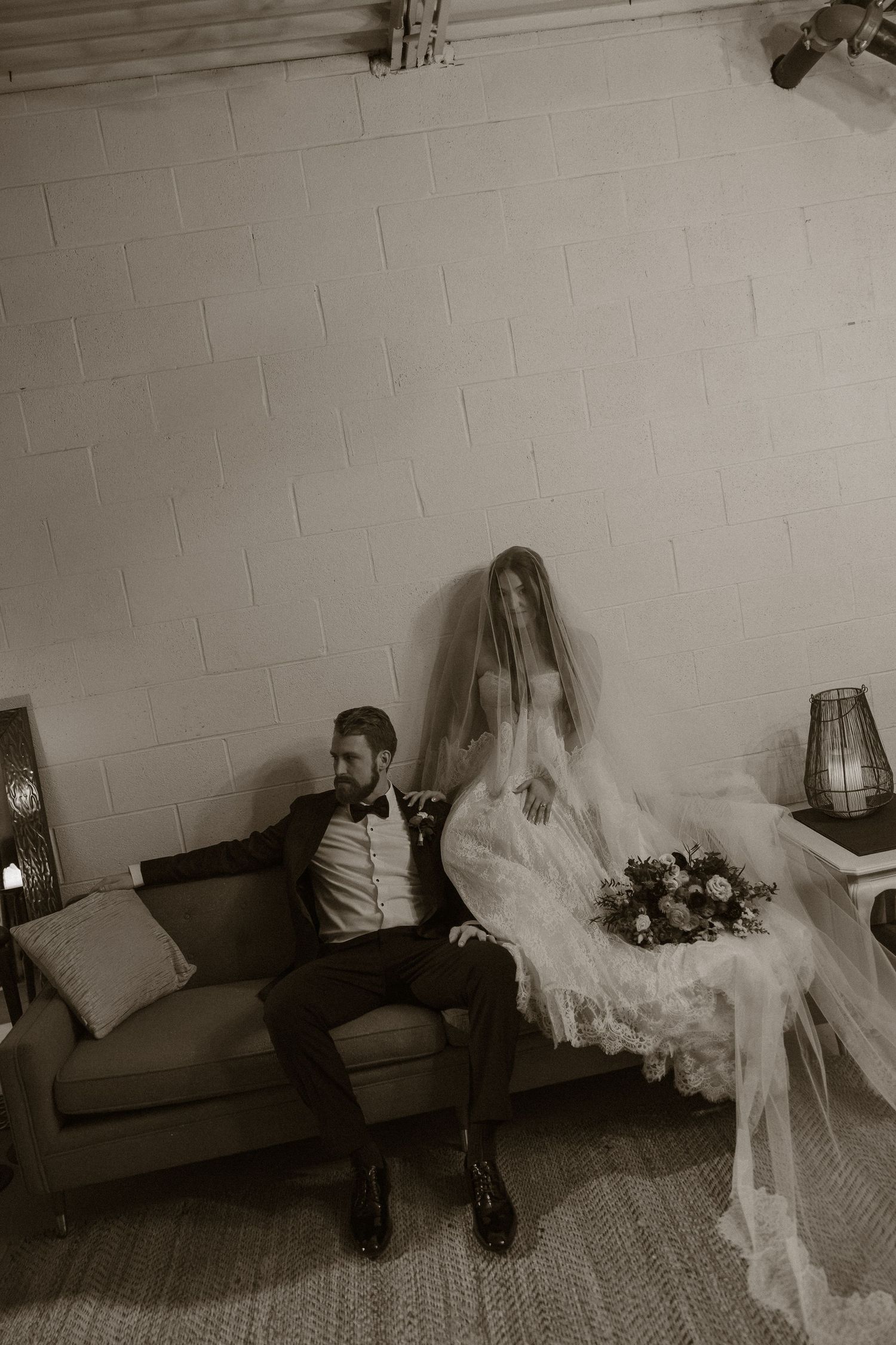 A black and white wedding photo showing a couple sitting on a couch, with the bride in a flowing veil and gown.