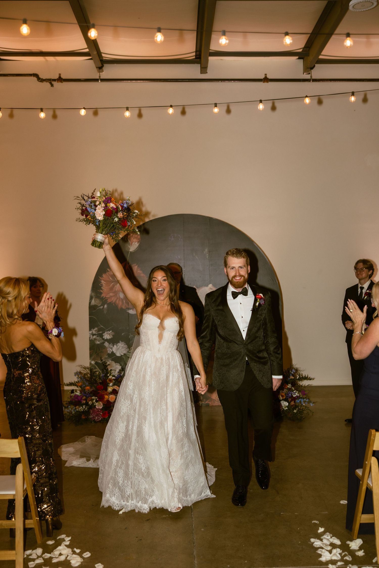 Wedding party dances and celebrates in a modern industrial venue with string lights overhead.