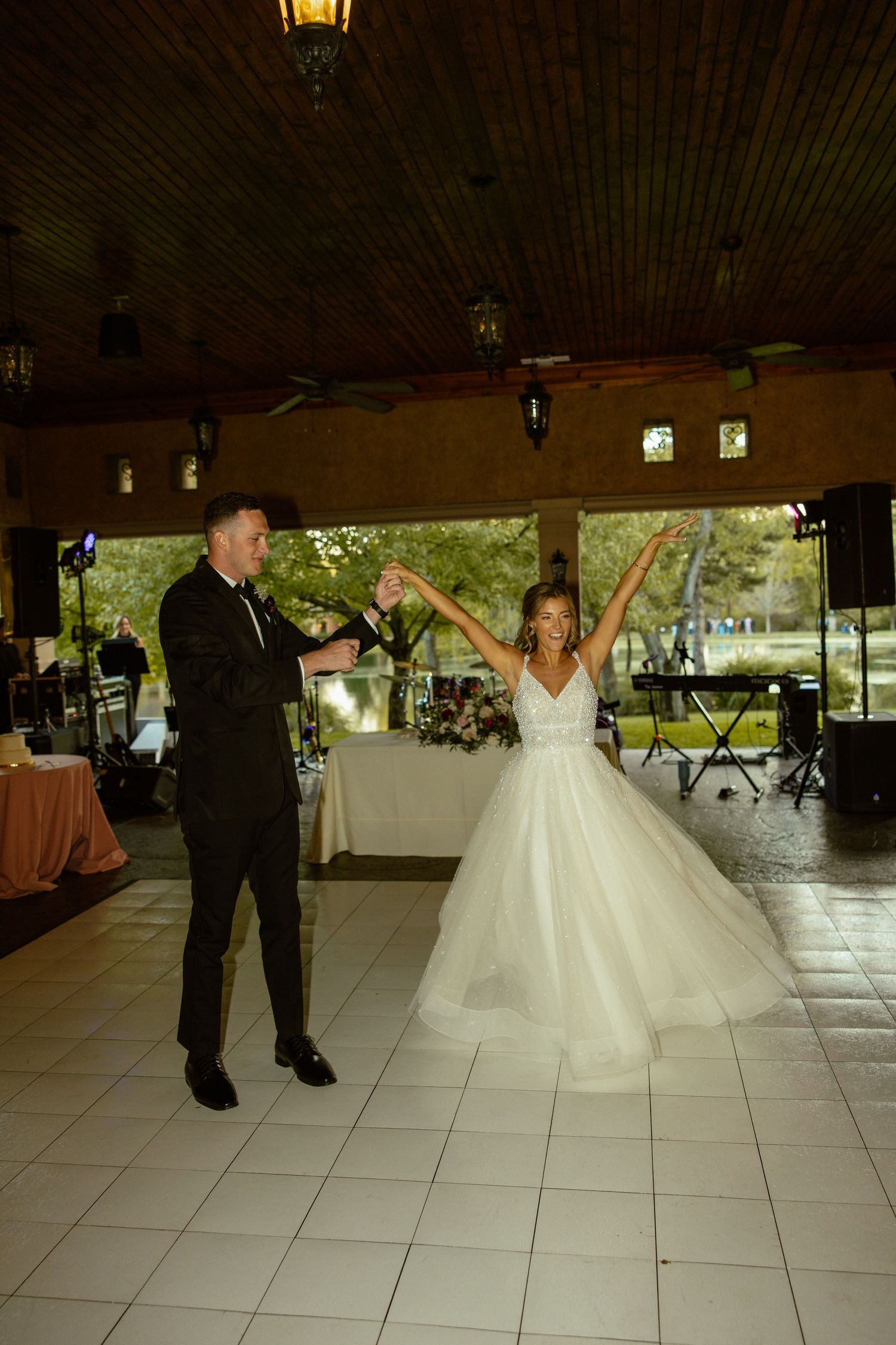 Wedding couple dances on white tile floor under covered pavilion with warm lighting.