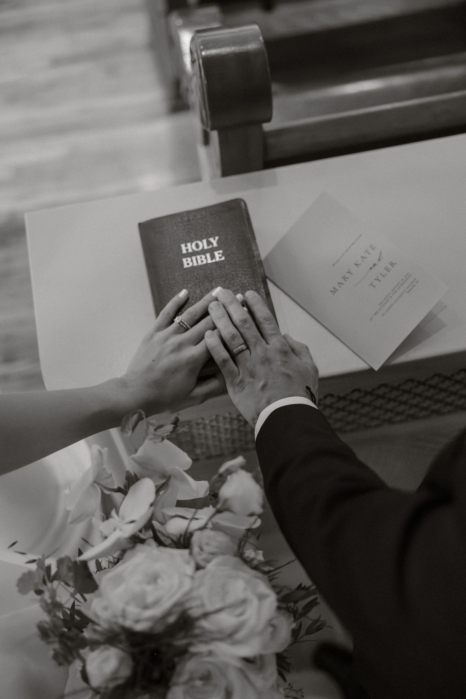 Black and white detail shot of hands holding a bible with wedding flowers on a desk.