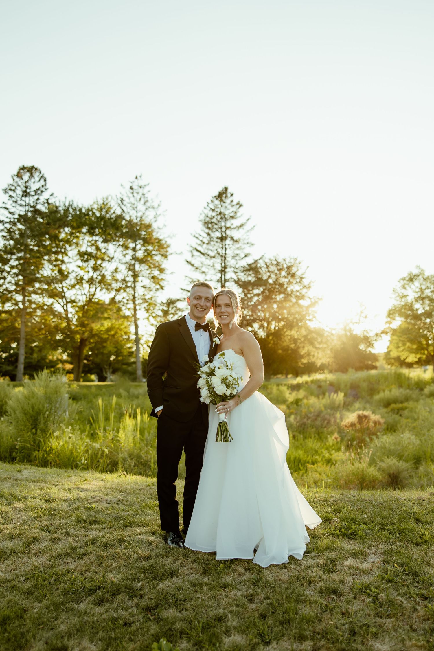 Wedding couple poses in a sunlit field at golden hour with tall trees in the background.