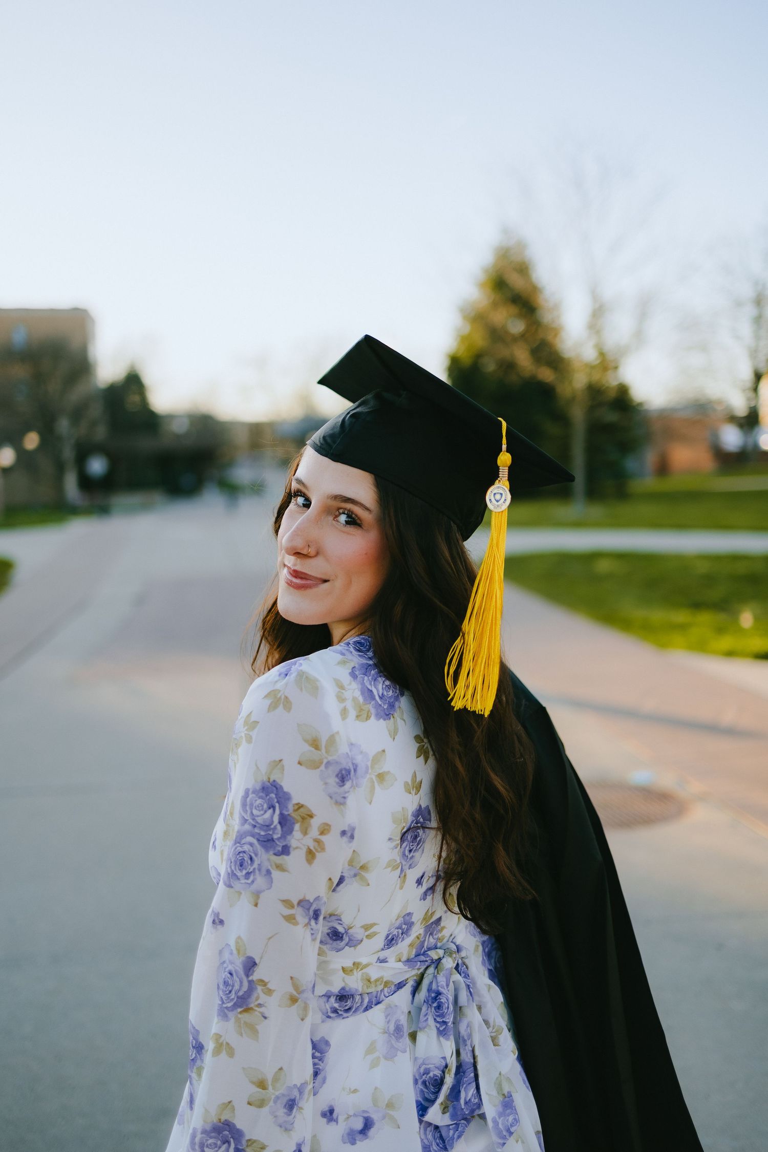 A graduate in a black cap and gown with a floral dress poses on a campus pathway at sunset.