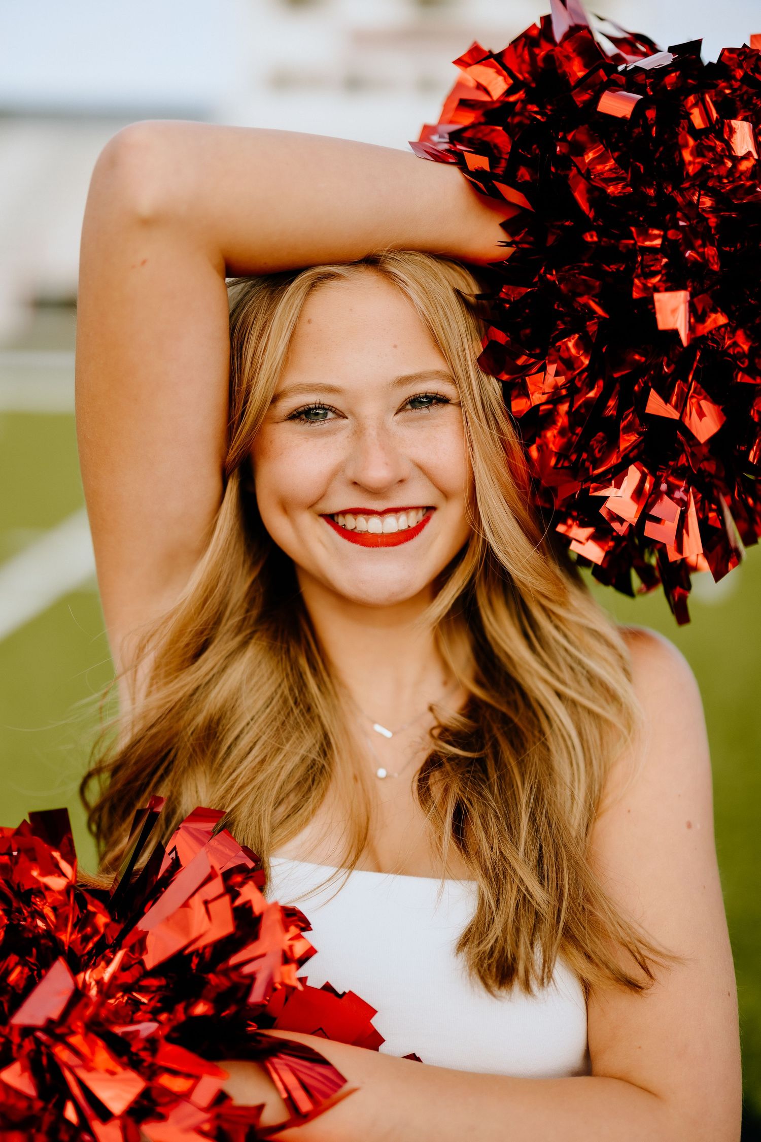 Cheerleader poses with red pom poms in white uniform on sports field.