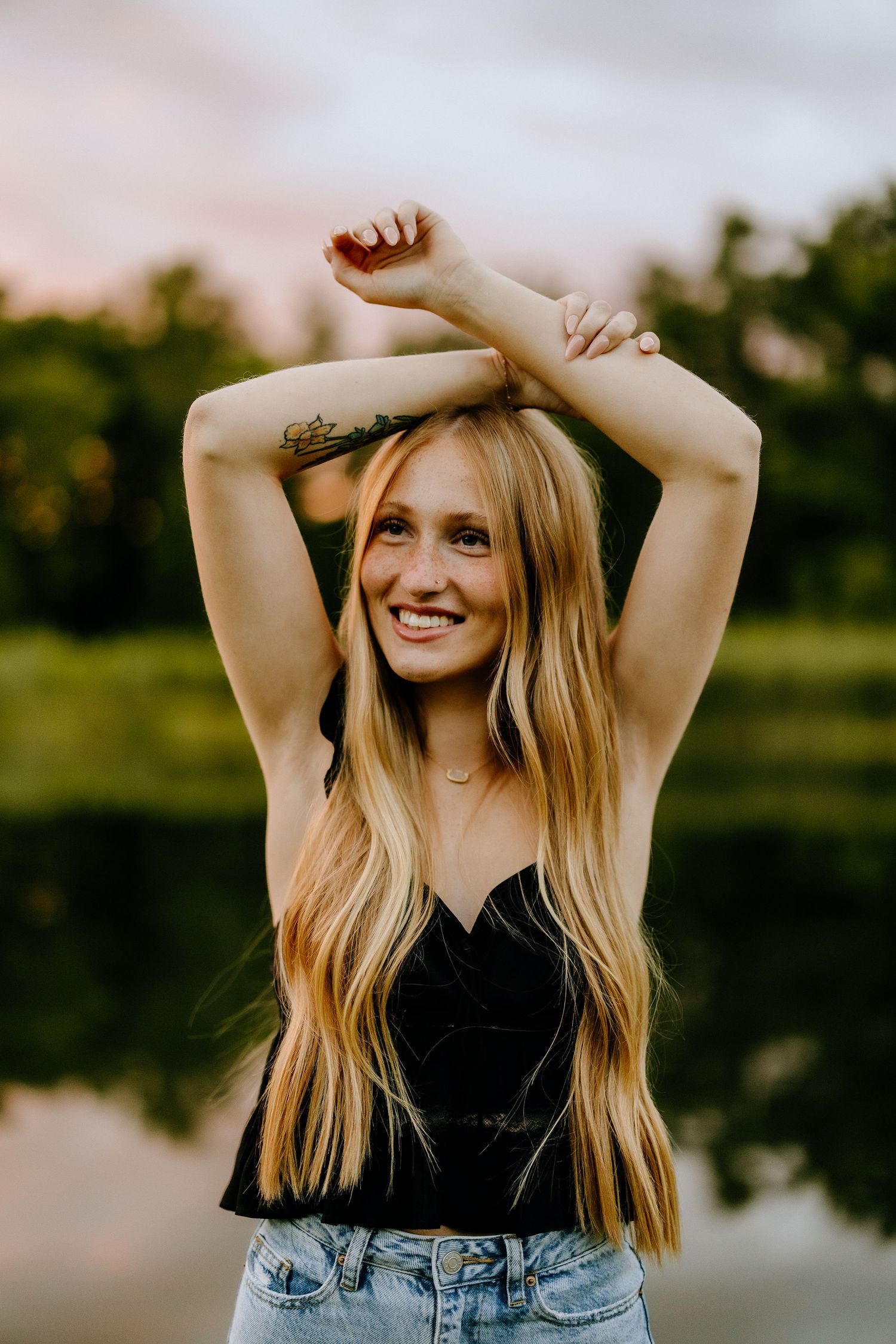 Person in black top and jeans poses playfully with arms raised during sunset photoshoot.