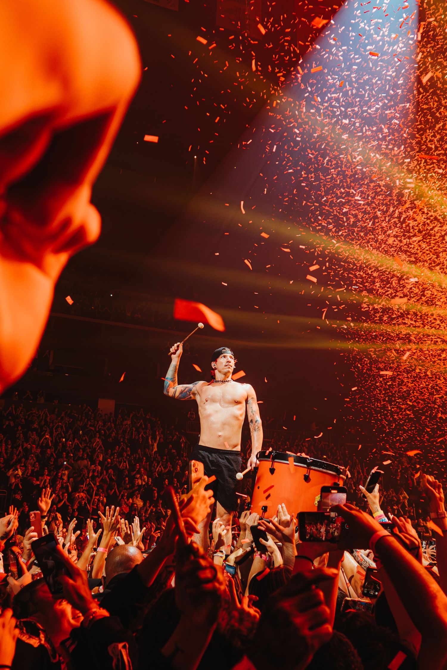 Concert performer stands shirtless on stage with crowd and orange lighting effects.
