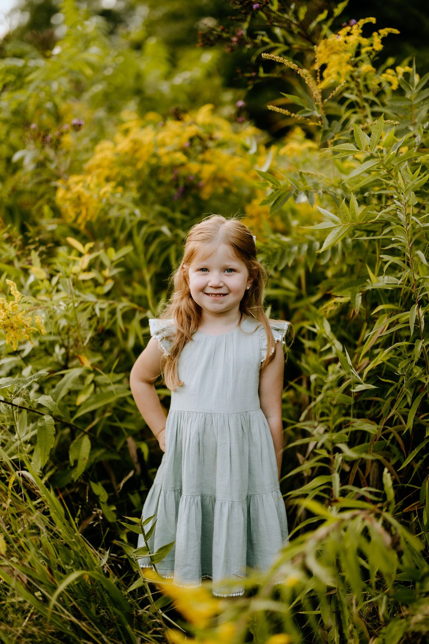 Young child in tiered dress stands among yellow flowering plants.