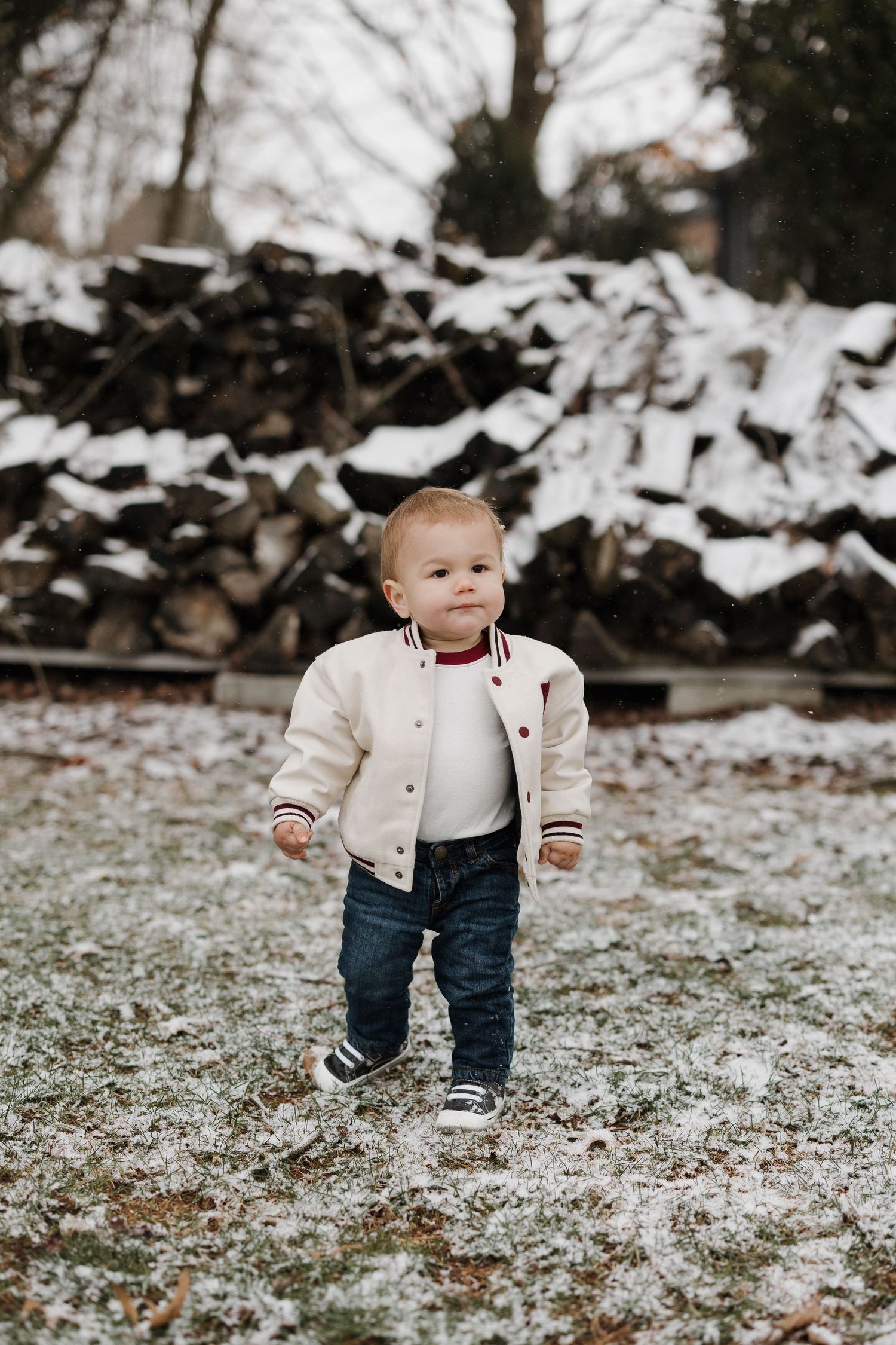Small child wearing cream jacket and jeans stands in snowy winter landscape with woodpile in background.