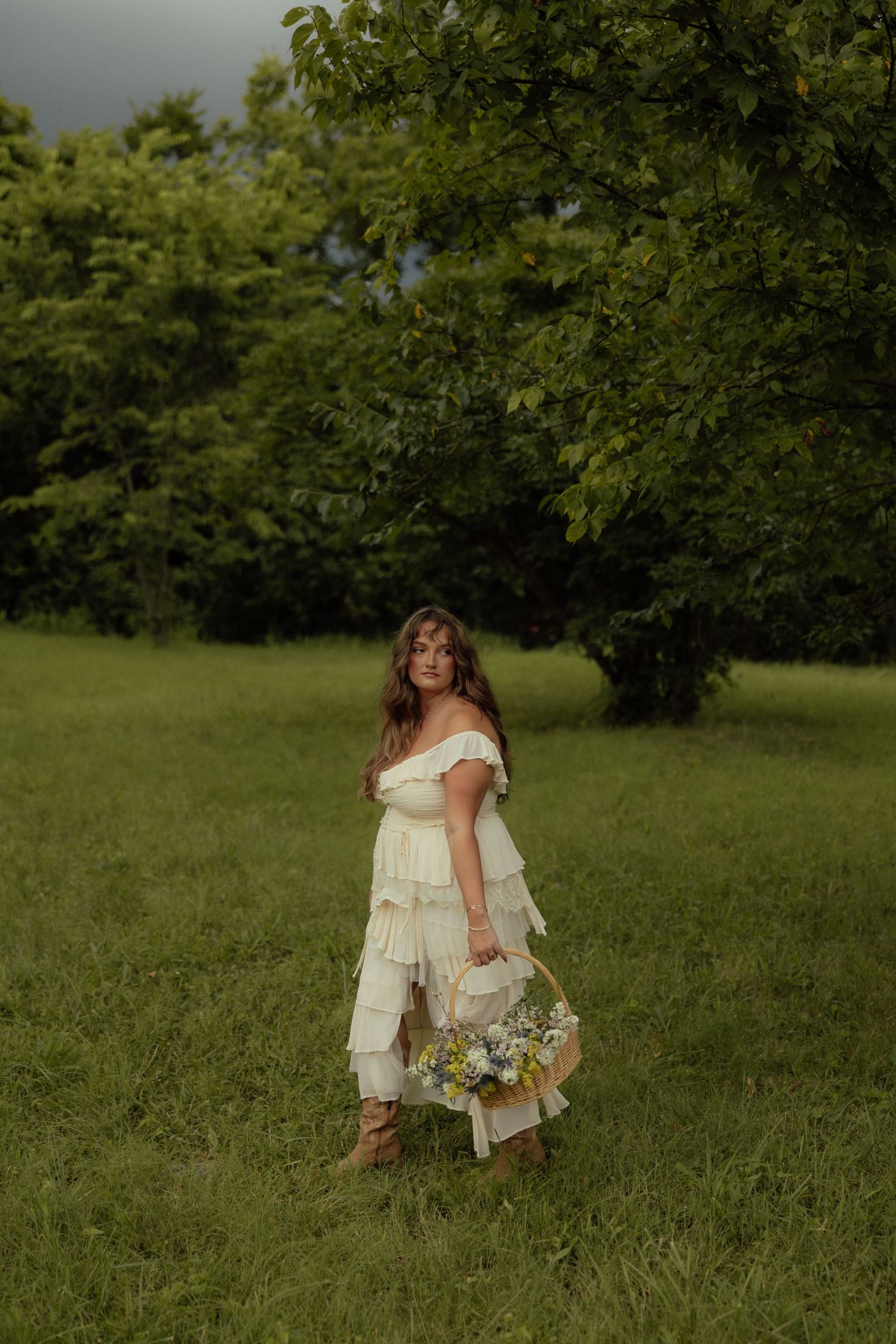 A dreamy outdoor photoshoot in a grassy field with someone wearing a flowing cream-colored dress holding wildflowers.