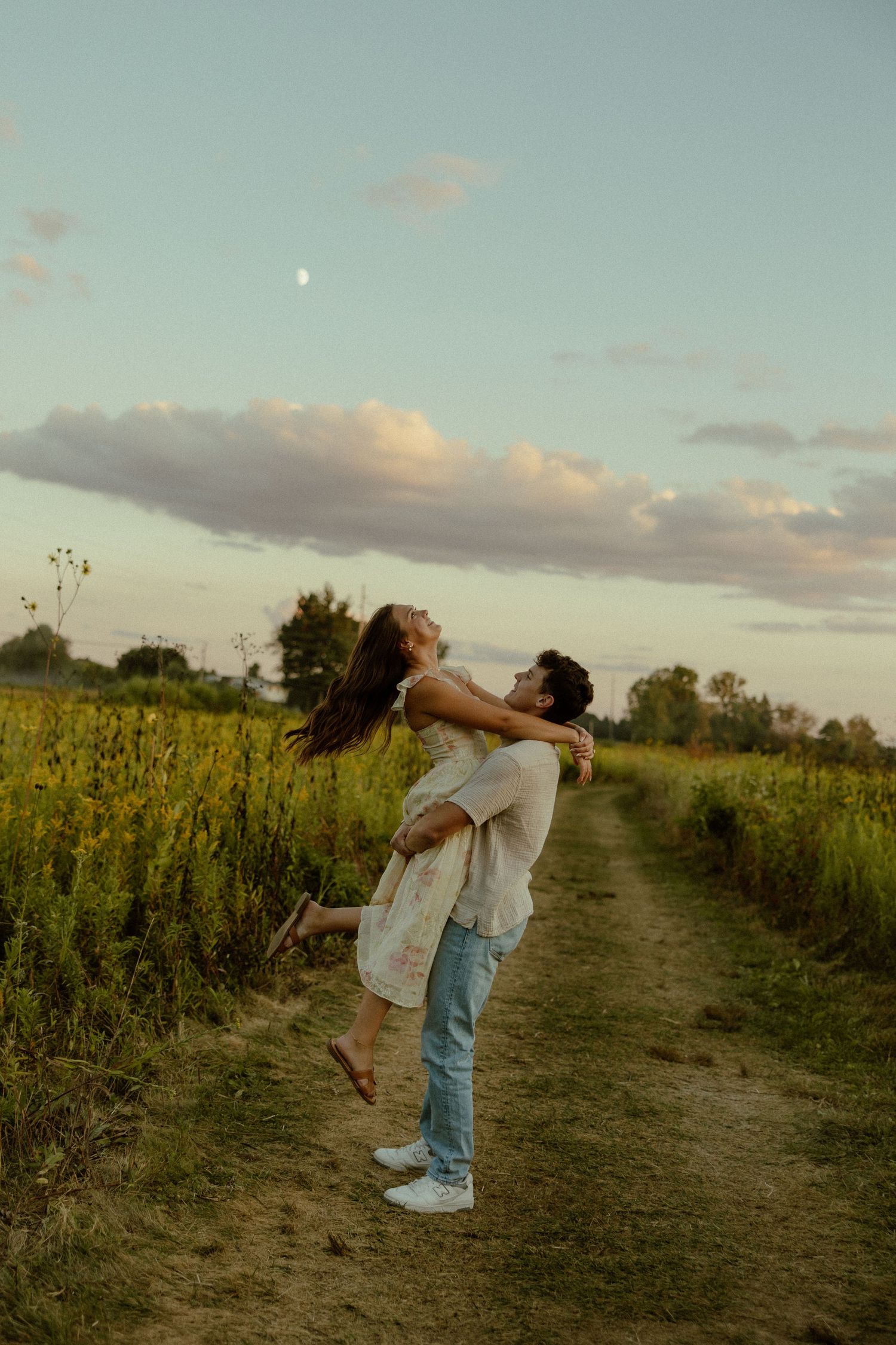 A couple embraces and leans back together on a dirt path through a golden field at sunset.