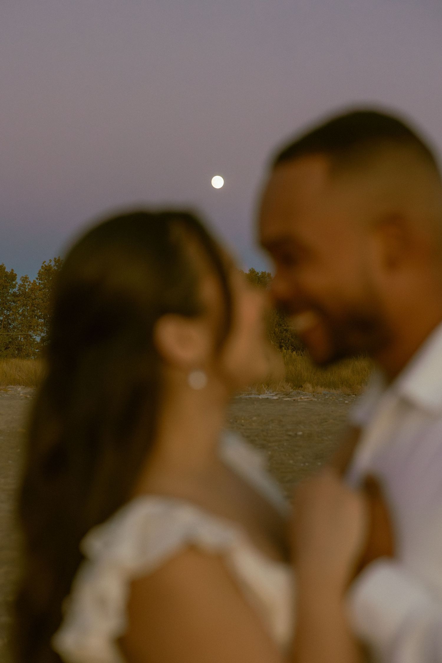 Romantic couple embraces and kisses at sunset by the water with moon visible in dusky sky.
