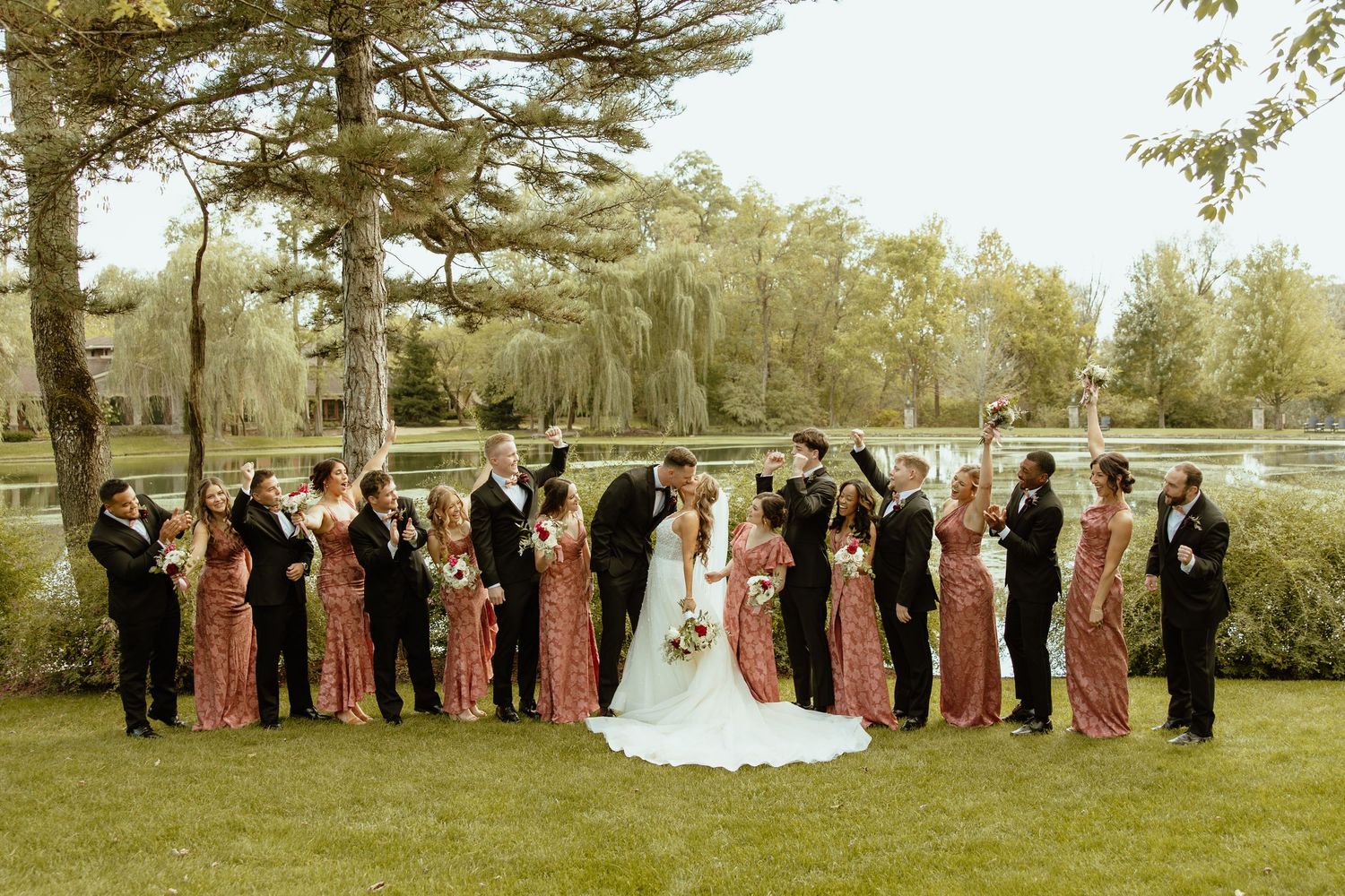 Wedding party poses in a line on lush green lawn with trees in the background during an outdoor celebration.