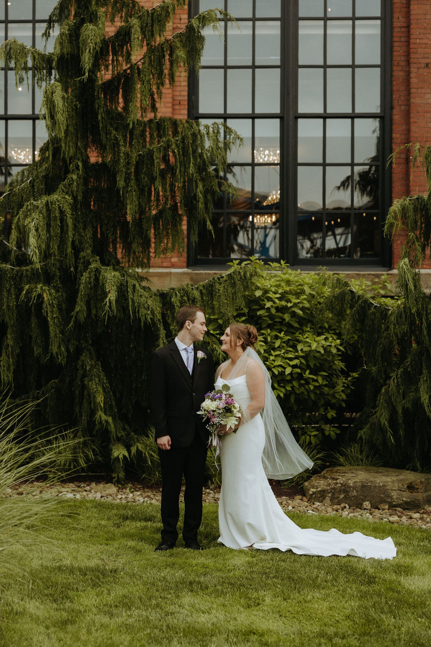 A wedding couple poses for portraits in front of a historic brick building with hanging willow trees.