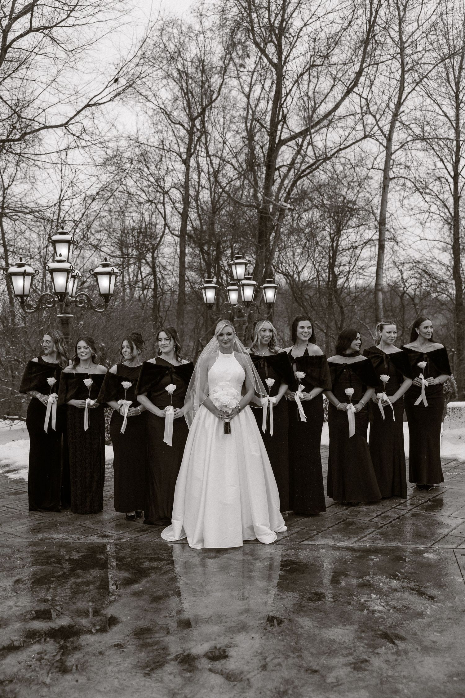 A wedding party in black and white attire poses outdoors in winter with bare trees and vintage lanterns in the background.
