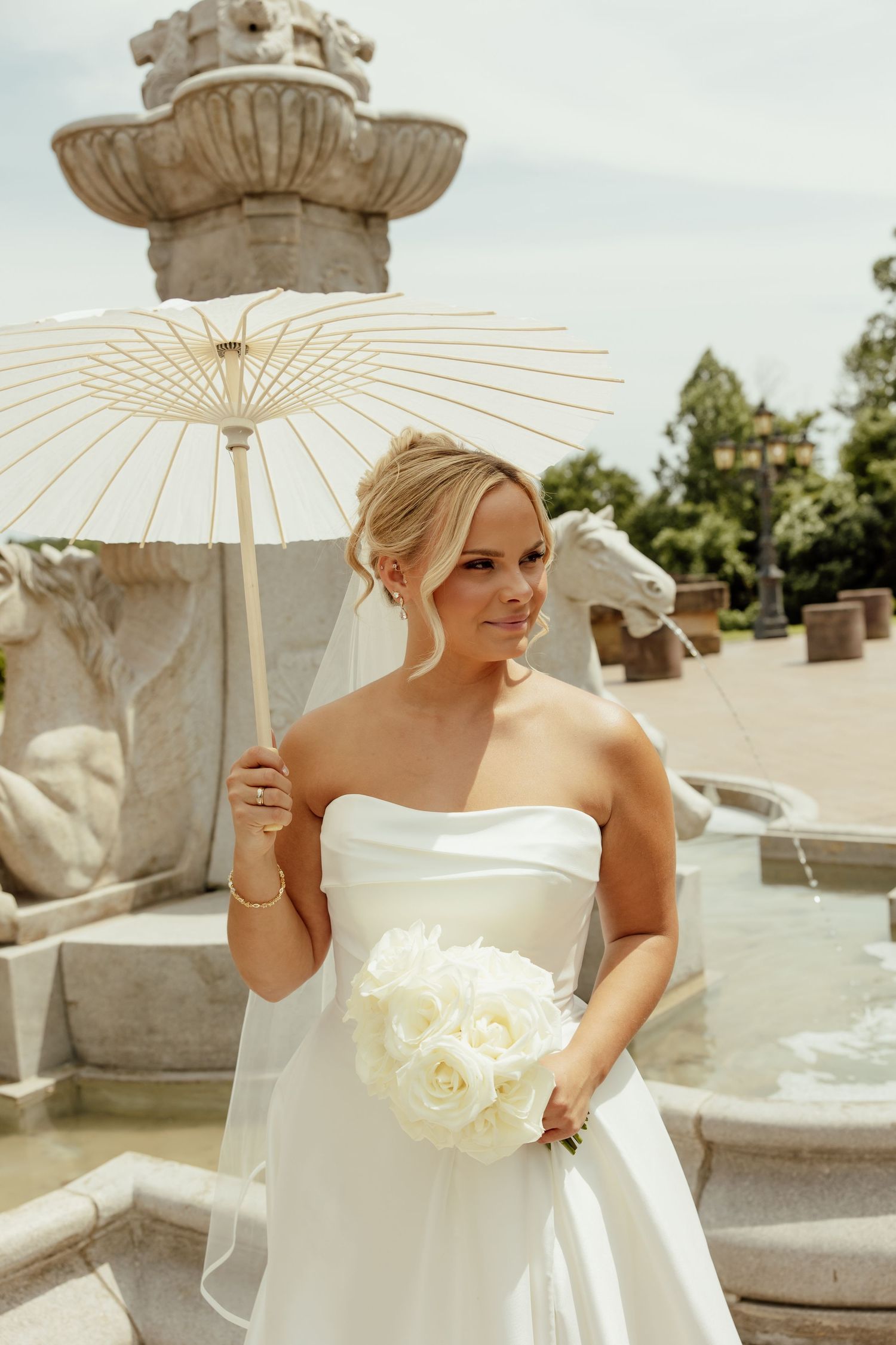 Elegant bride in white strapless gown holds parasol and white rose bouquet near ornate stone fountain on sunny day.