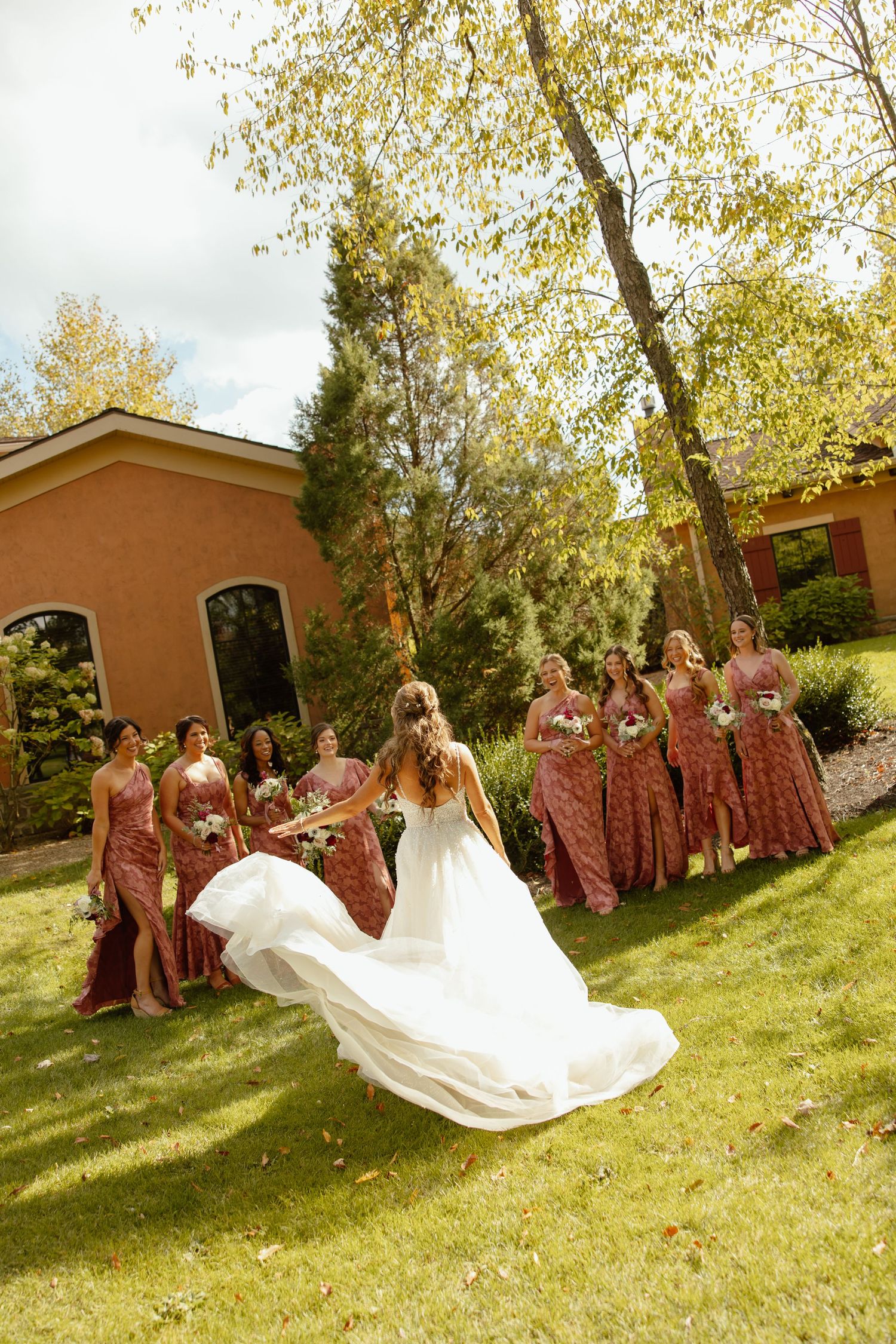 Bridesmaids in burgundy dresses help with a flowing white wedding gown train on a sunny lawn.
