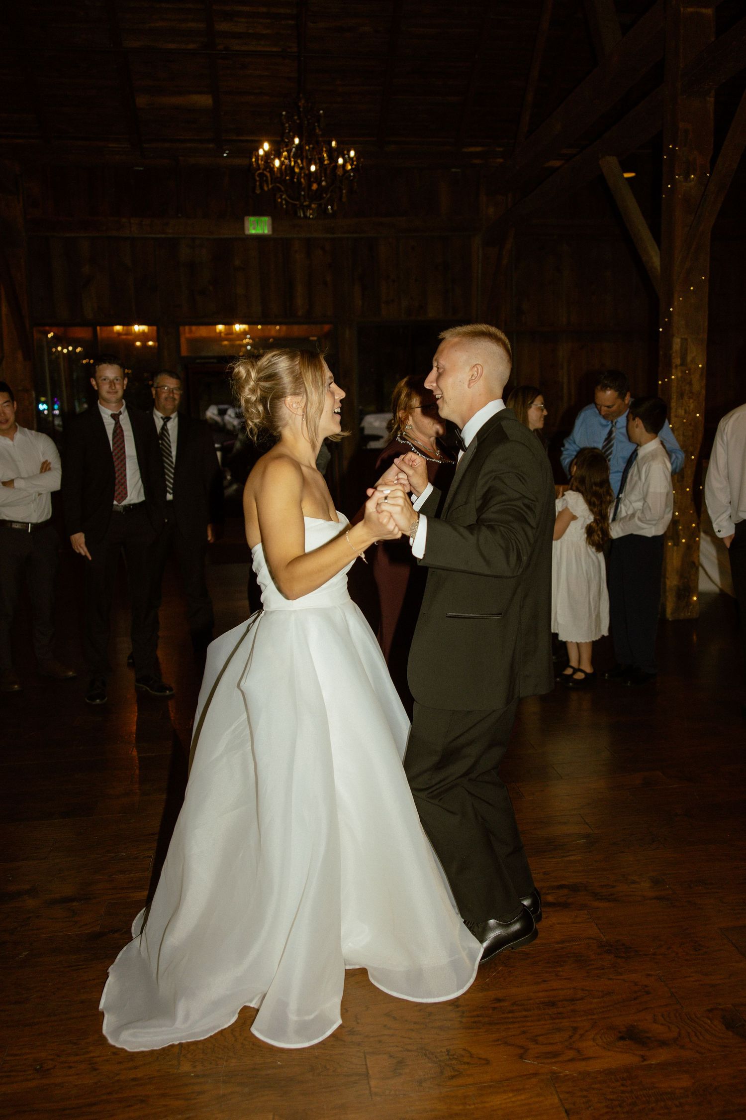 A couple dances together on a wooden floor during a wedding reception in a rustic venue with warm lighting.