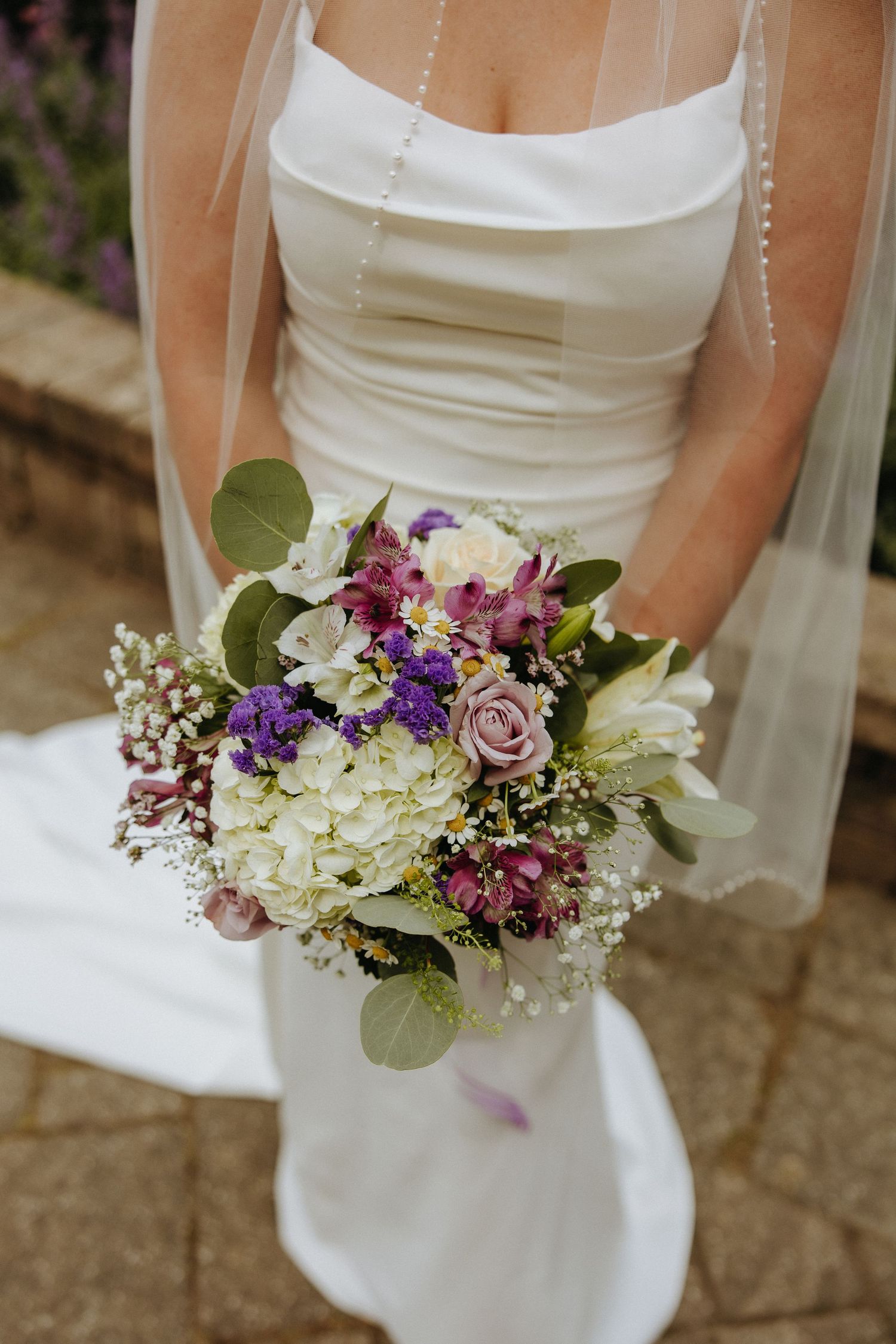 Beautiful wedding bouquet with white hydrangeas, purple flowers, and greenery held against a white wedding dress.