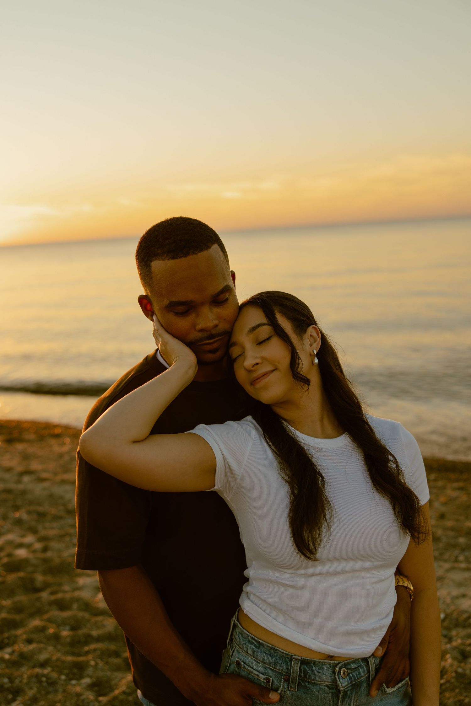 A couple shares an intimate moment on the beach at sunset with ocean waves in the background.