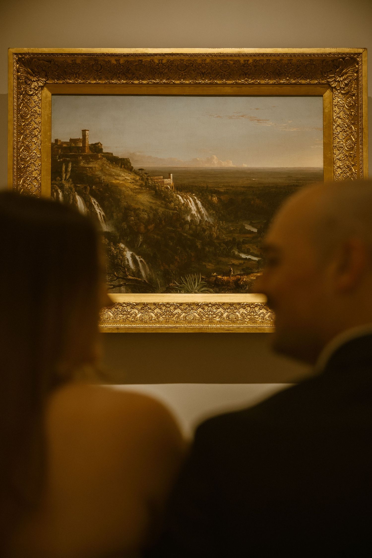 Museum visitors observe framed landscape paintings displayed in ornate gold frames on a gallery wall.