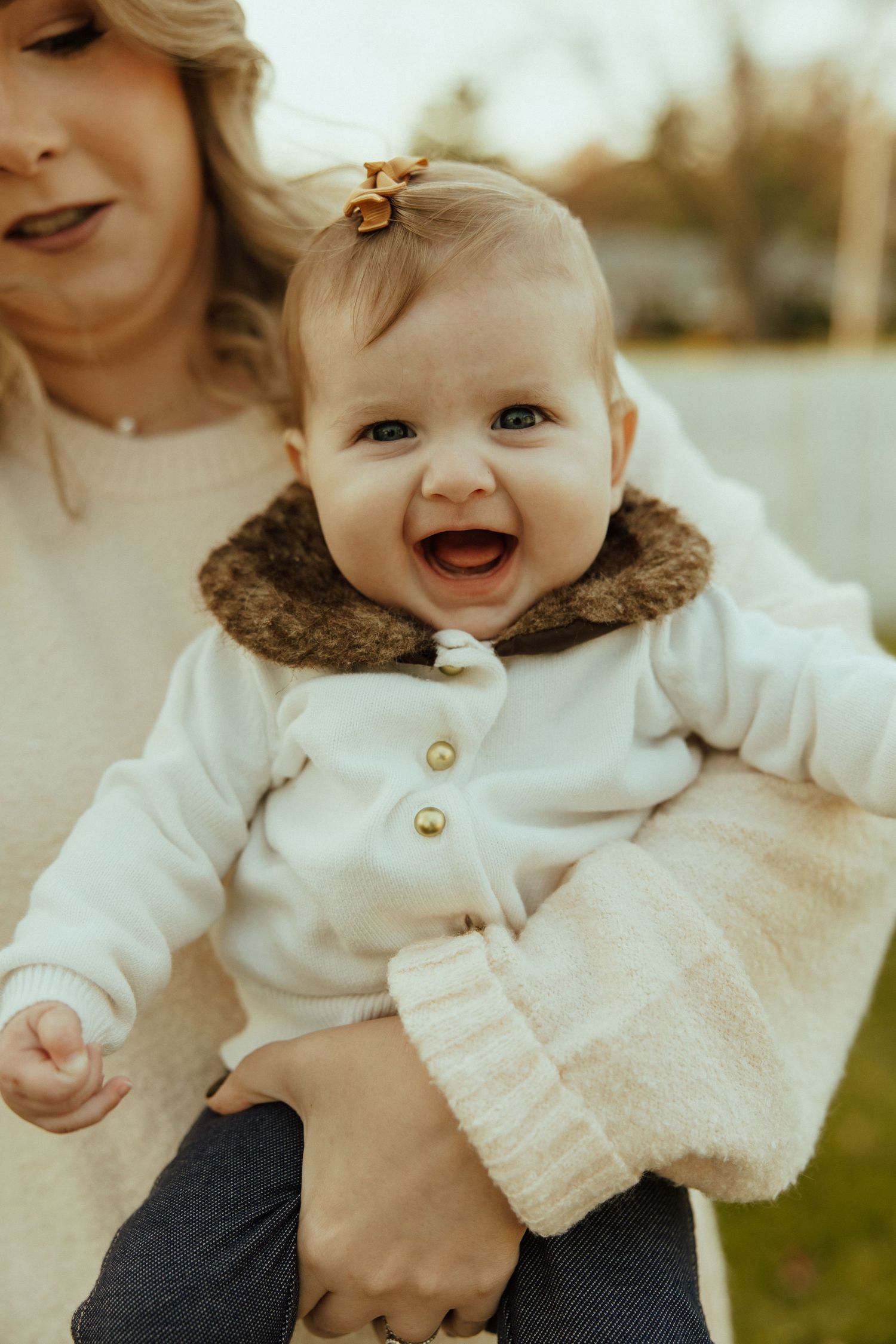 Baby in white sweater smiles joyfully while being held outdoors in warm sunlight.