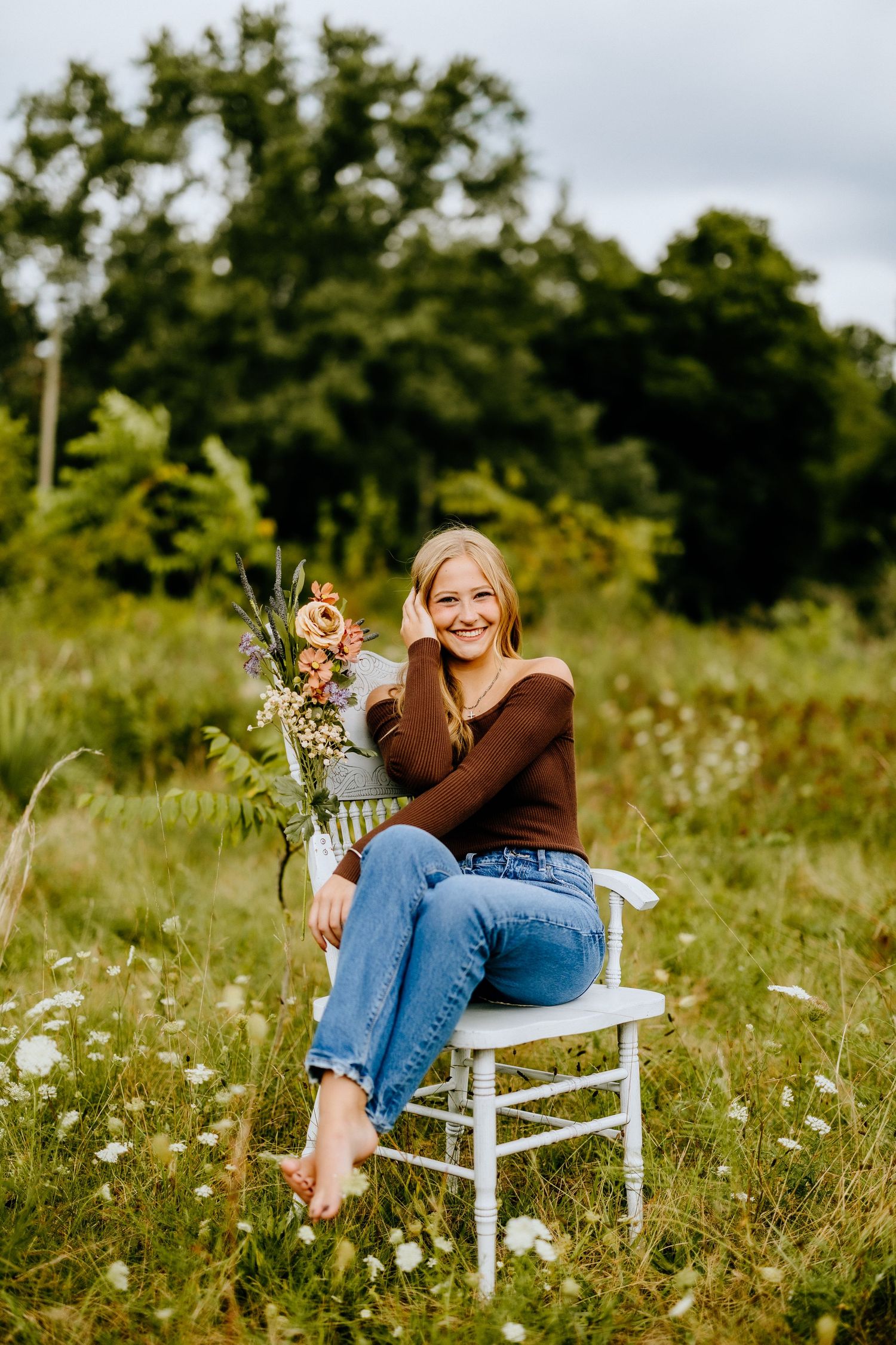 Person in brown sweater sits on white chair in meadow with flower arrangement.