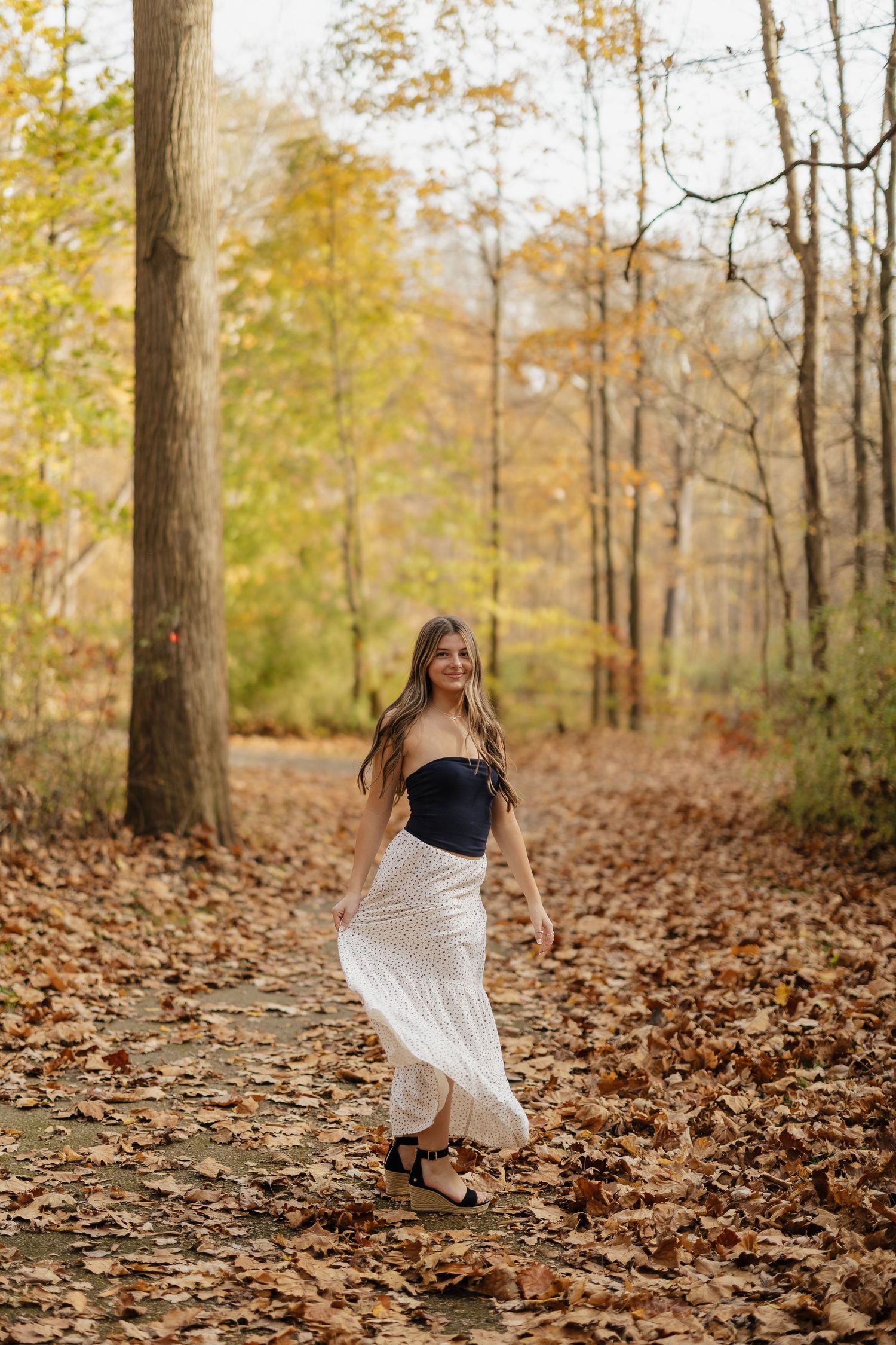 A flowing white maxi skirt catches movement among fallen autumn leaves on a forest path.