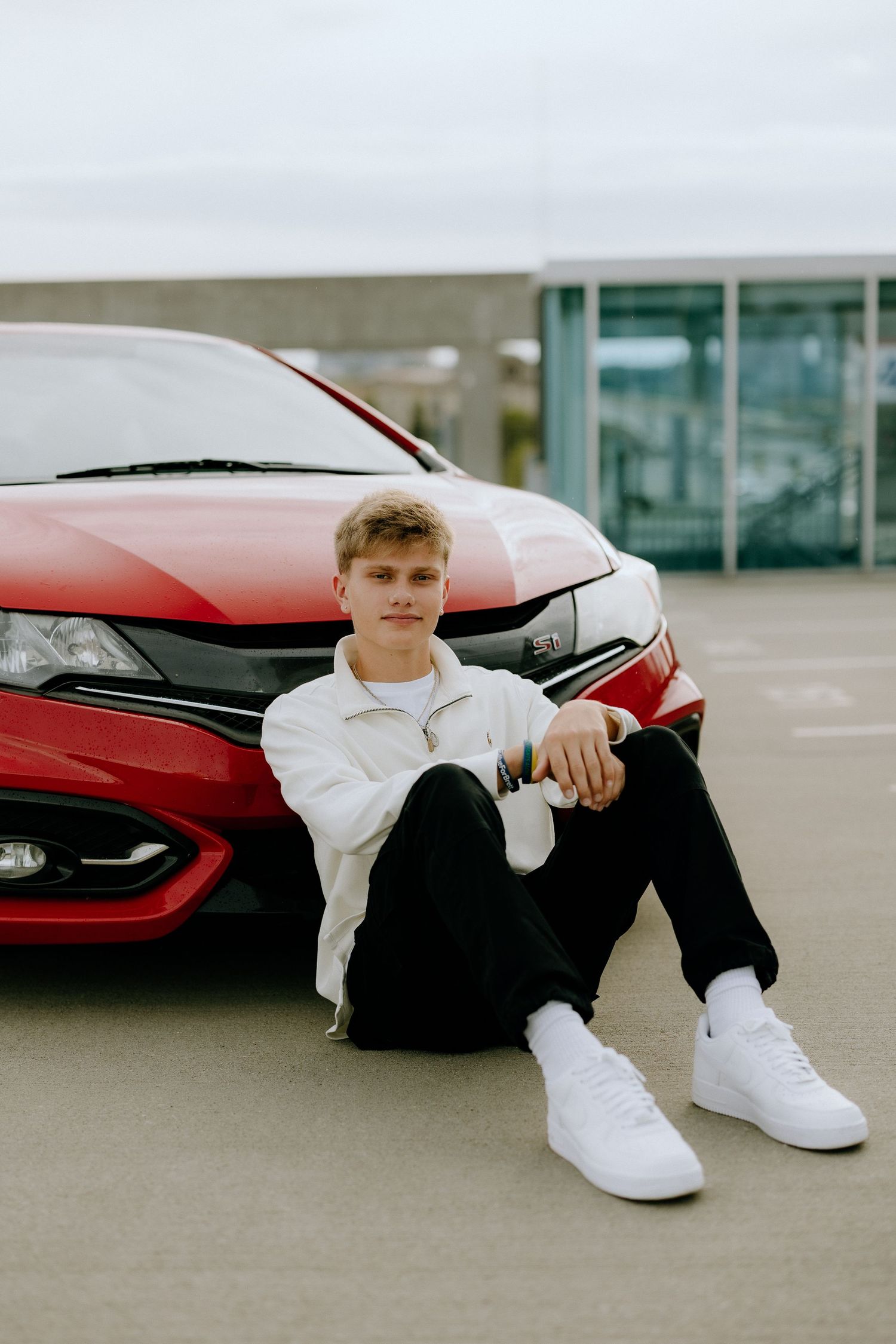 Urban fashion portrait sitting against red car wearing white sweater.