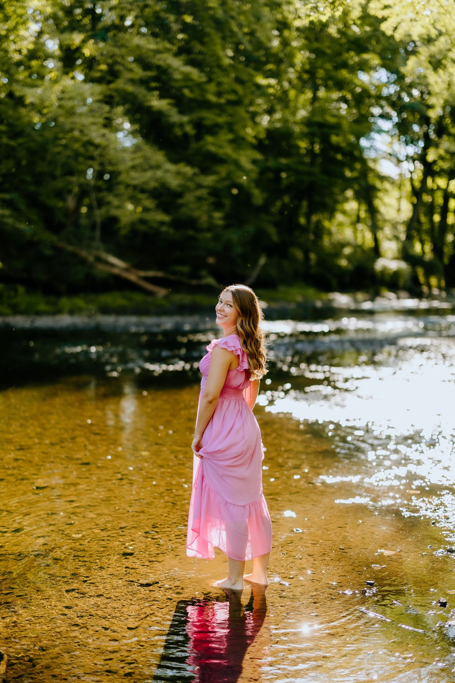 A flowing pink dress reflects in shallow stream water surrounded by green forest.