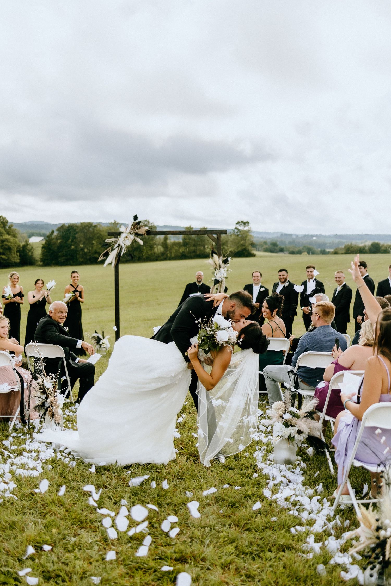 An outdoor wedding ceremony in a scenic countryside field with white flower petals scattered along the aisle at sunset.