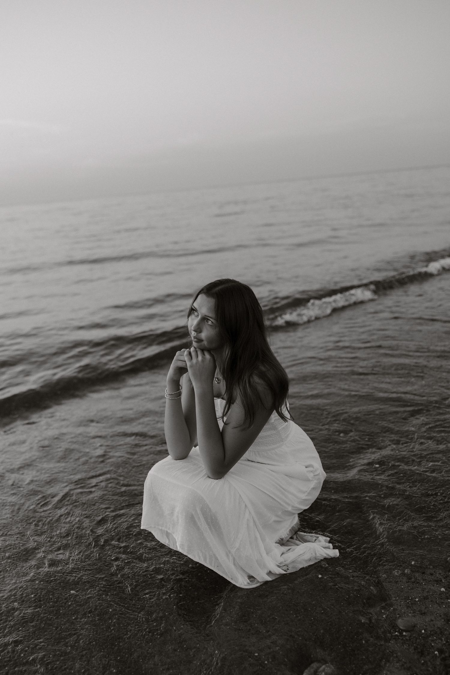 A person in a white dress sits contemplatively by the ocean shore in black and white photography.