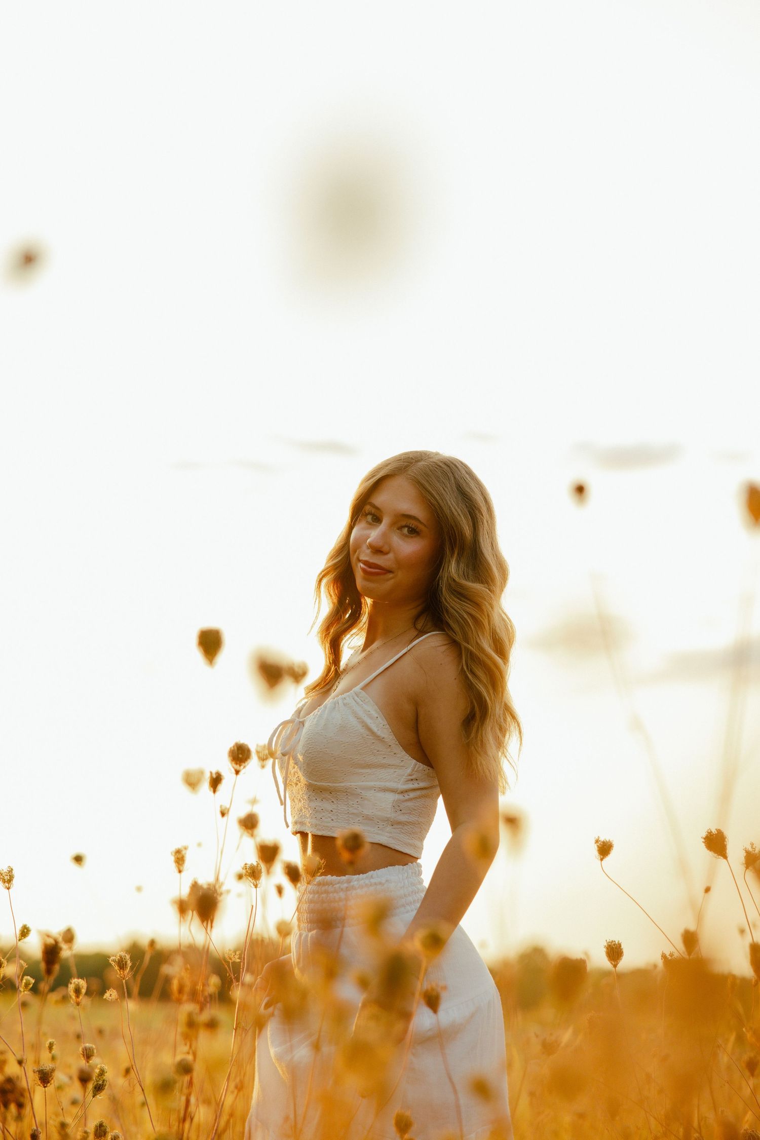 Person in white outfit poses in golden wildflower field at sunset with warm backlighting creating dreamy atmosphere.