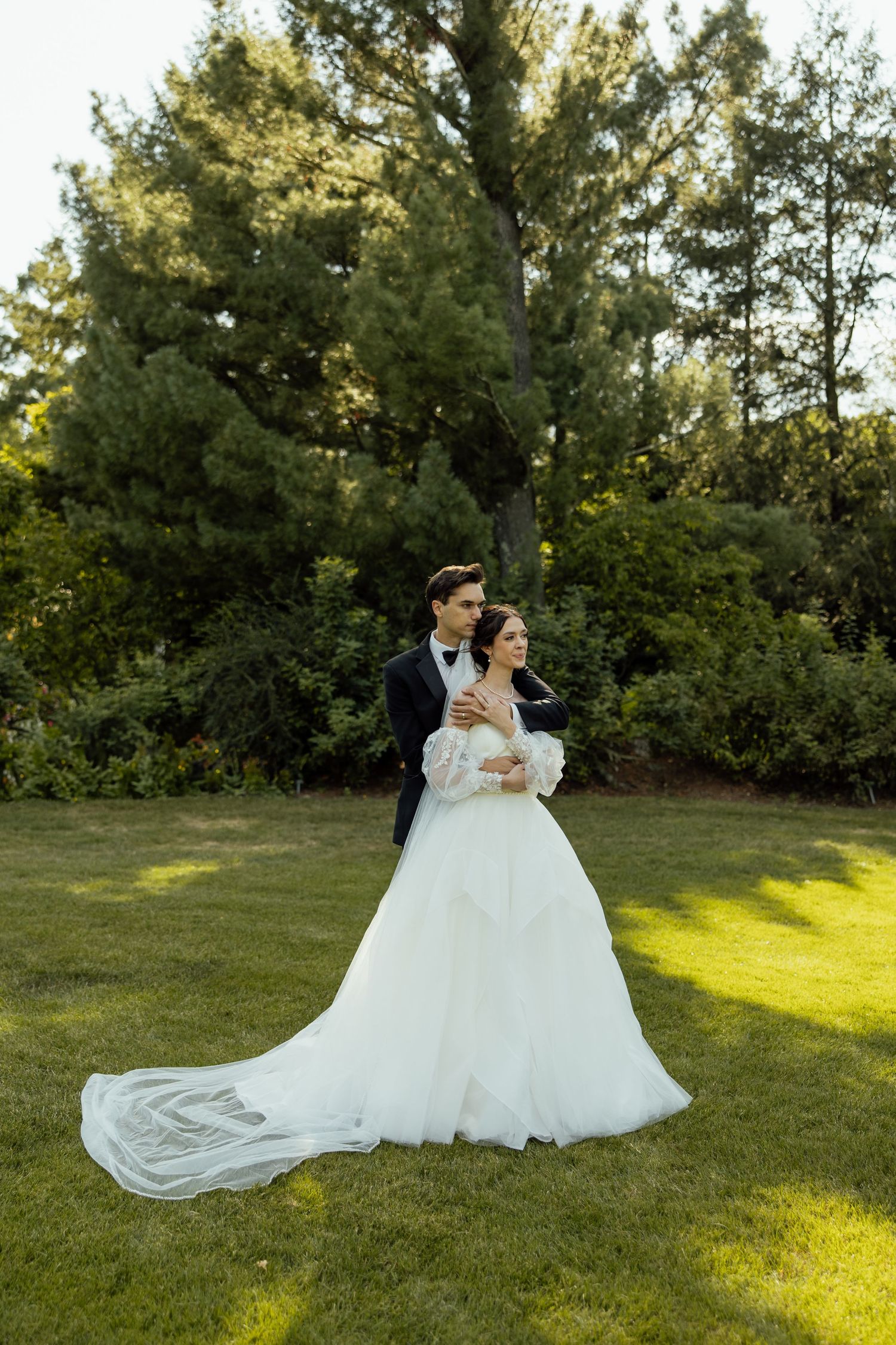 A couple in a white wedding dress and suit embrace on a sunlit lawn surrounded by evergreen trees.