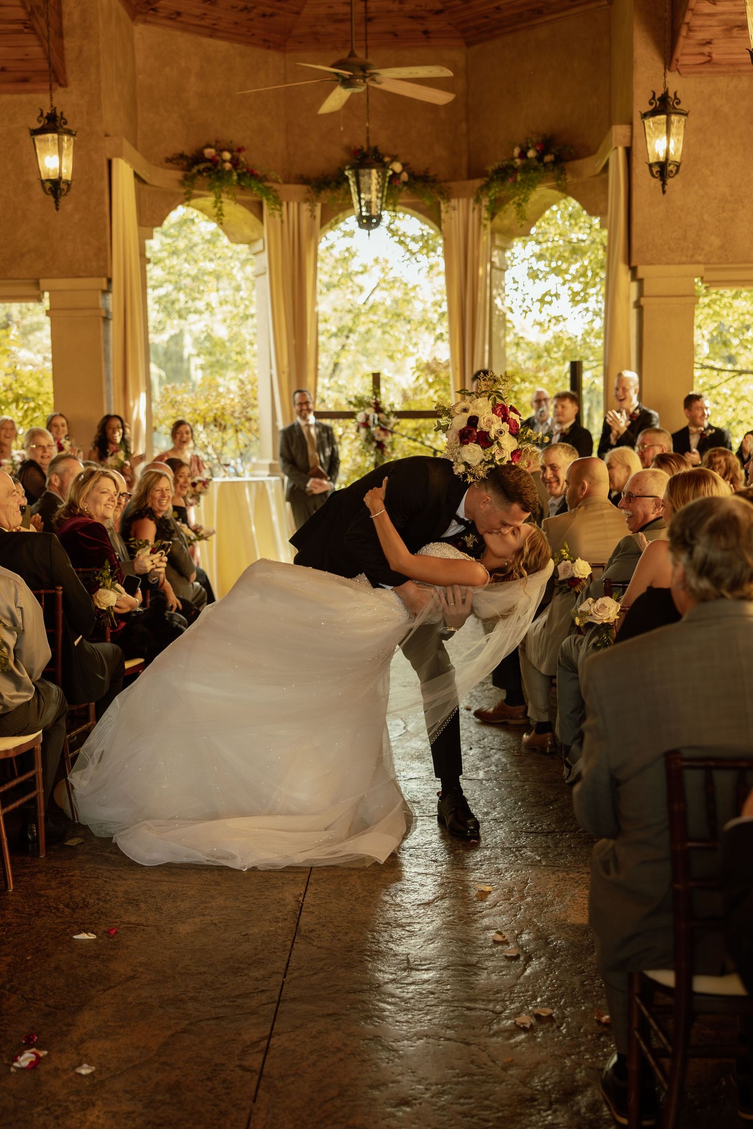 Couple shares romantic dip kiss during wedding ceremony in elegant outdoor pavilion with hanging lanterns.