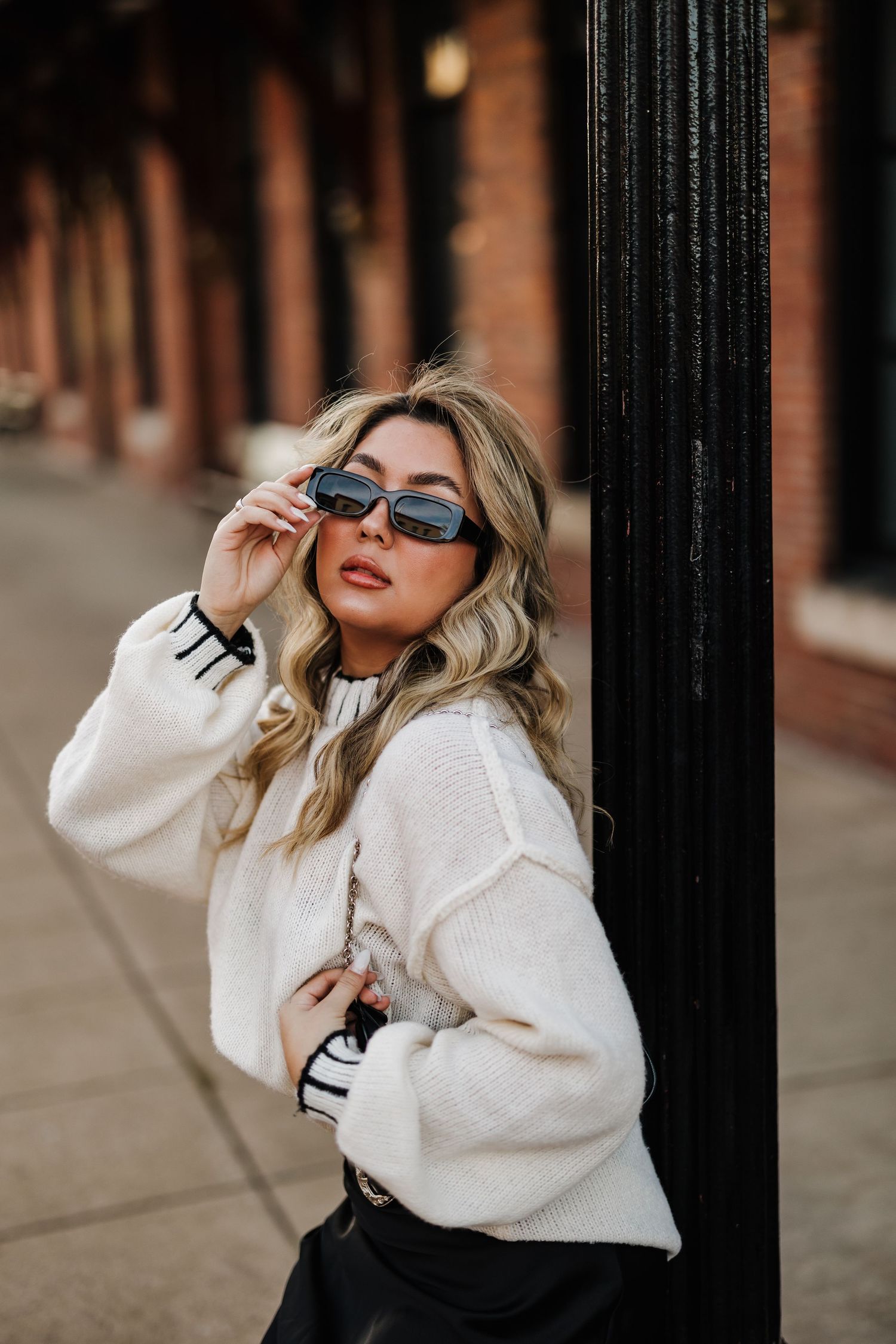 Street fashion photo featuring a cream sweater and sunglasses against brick architecture.