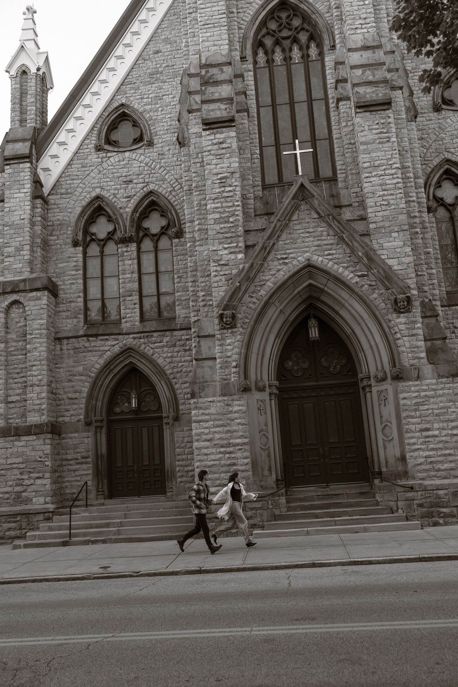 Two people run past the Gothic stone facade of a historic church with arched windows and doorways in black and white.