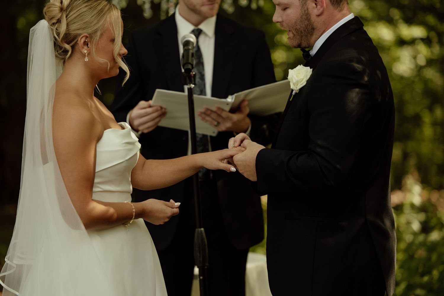 A couple exchanges rings during an outdoor wedding ceremony surrounded by greenery and natural light.