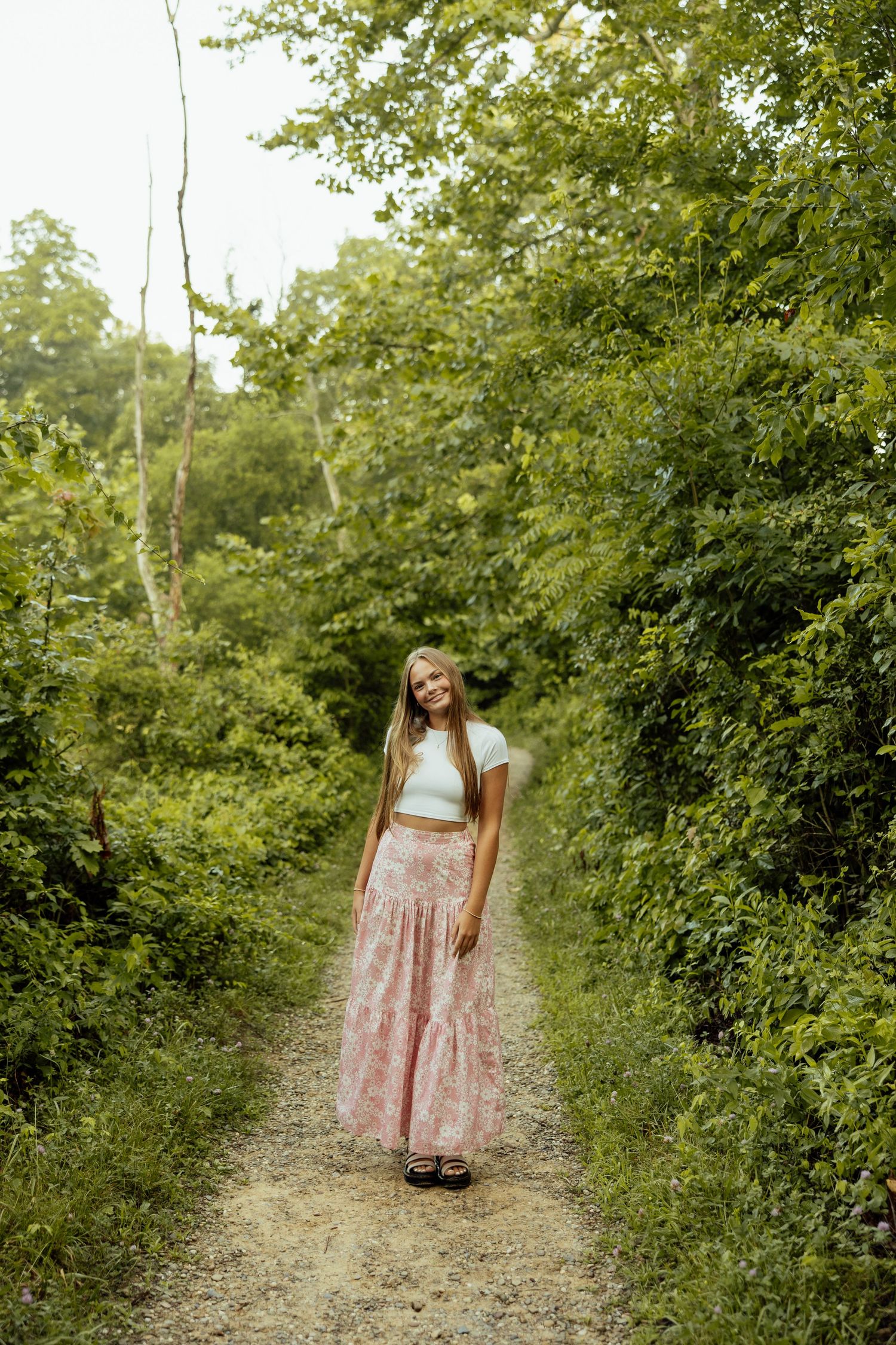 A person in a floral dress and mint top walks along a winding dirt path surrounded by lush green foliage.