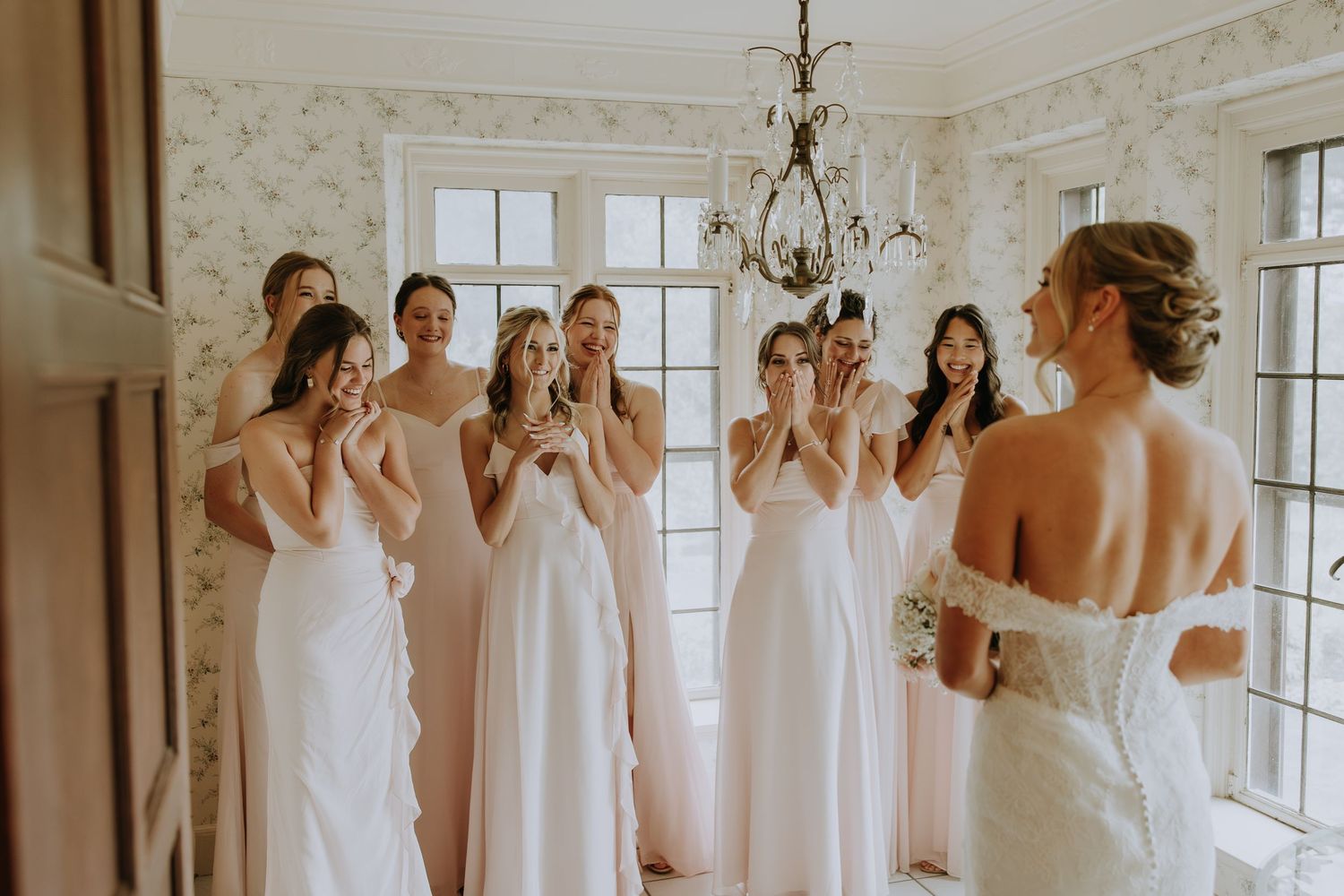 Bridesmaids in light colored dresses gather excitedly in a bright room with white walls and windows.