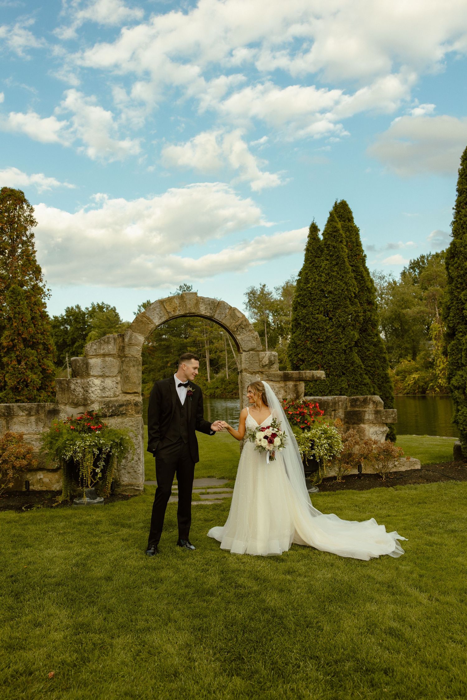 Wedding couple poses for photos in front of stone archway ruins on green grass under blue sky.