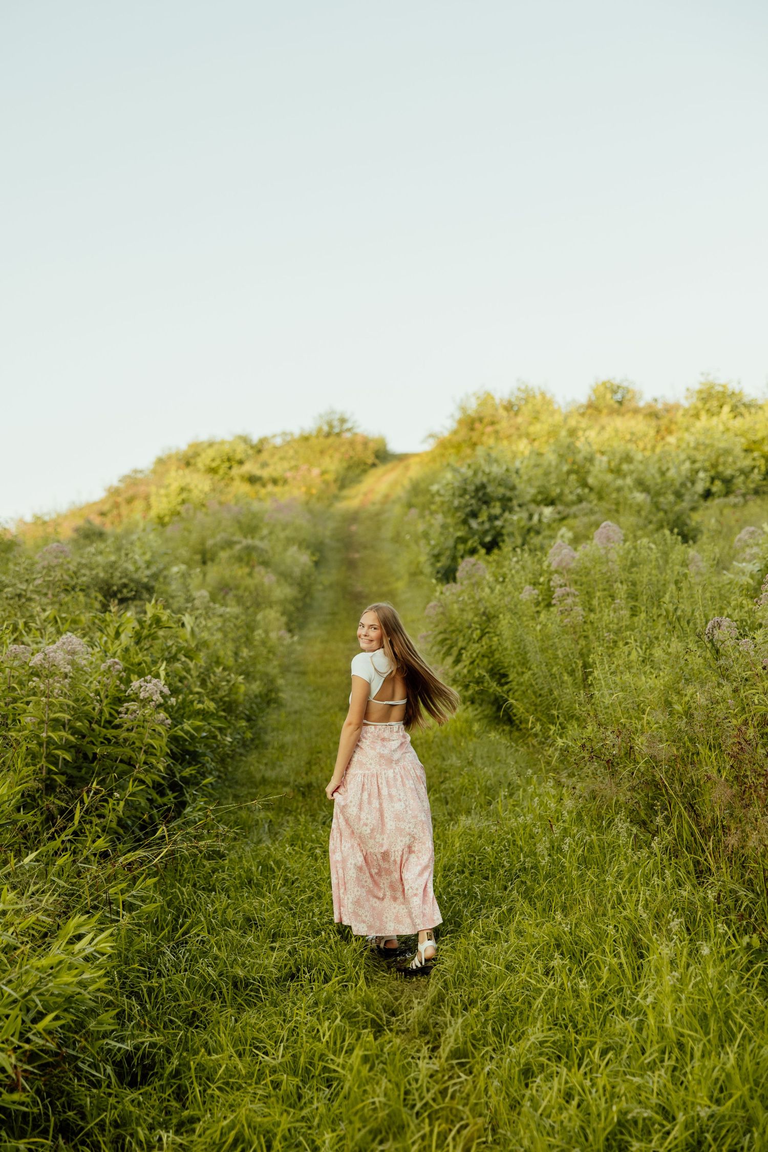 A person in a flowing pink dress walks up a grassy hillside path surrounded by lush summer foliage.