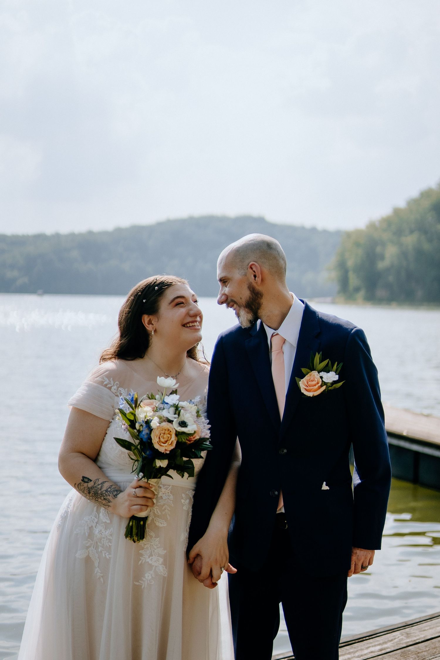 Joyful wedding couple laughing together on dock with lake and mountains in background.